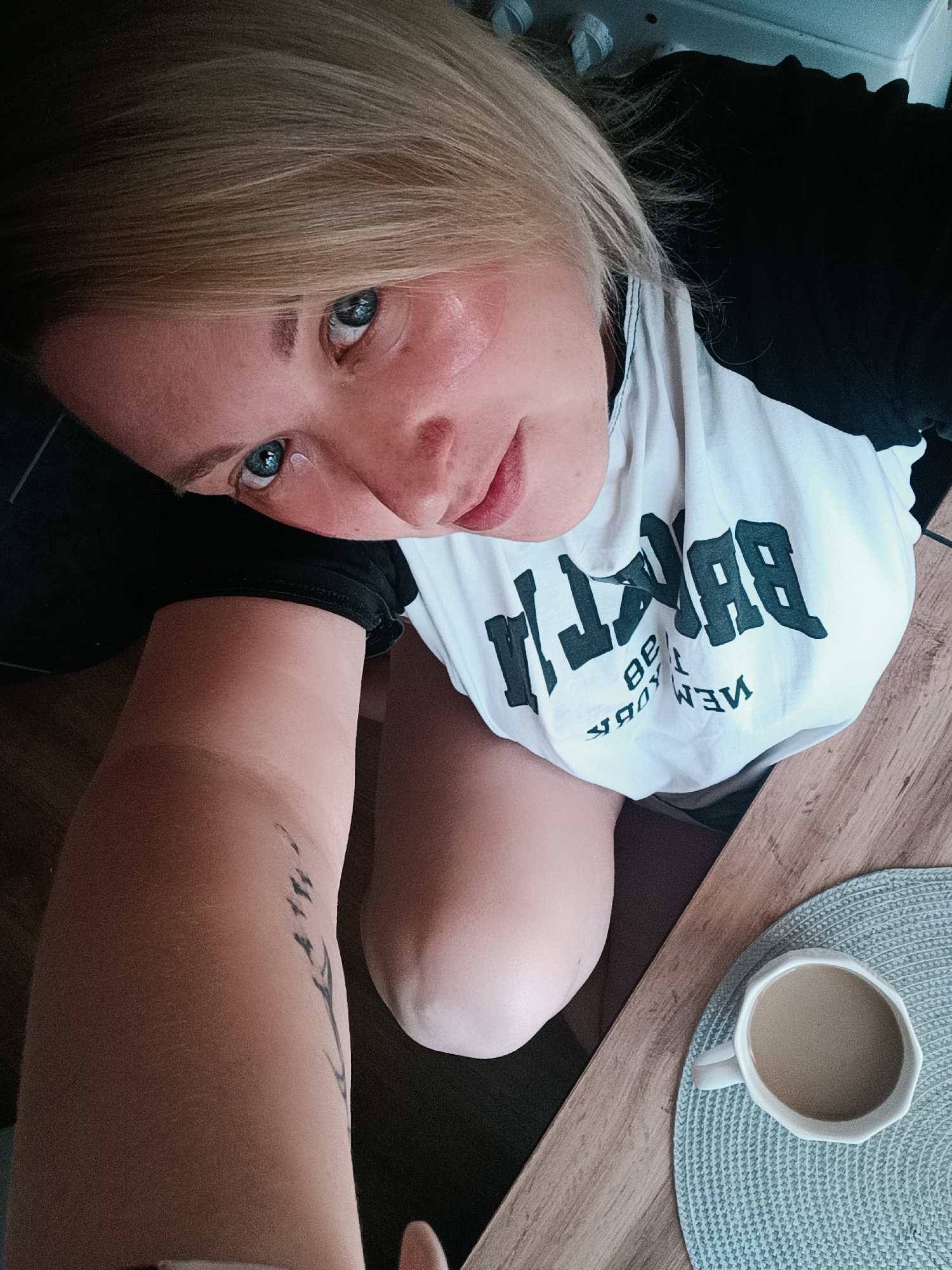 A woman with blonde hair and blue eyes is resting her head on her arm, which has a tattoo of a plant. She is wearing a white t-shirt with black sleeves and the word "BROOKLYN" printed on it in black letters. A mug of coffee rests on a blue placemat on a wooden table. The location is Chaikovsky, Russia.