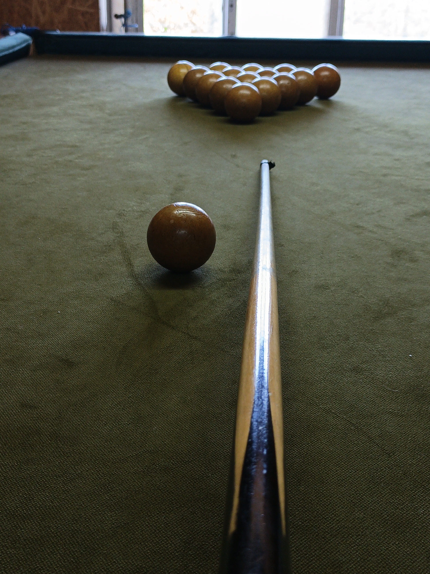 A low-angle, eye-level perspective captures a billiard game setup in an indoor setting located in Терновский округ, Ukraine. A billiard table, covered in a worn olive-green felt, dominates the frame. In the foreground, slightly left of center, a single solid brown ball rests on the felt. Extending from the bottom center of the frame and angled slightly towards the left is a wooden billiard cue stick, featuring a light wood shaft and a darker, stained grip section. Its tip is positioned to strike the brown ball. In the mid-ground, at the far end of the table and positioned directly in line with the cue stick, a complete rack of fifteen identical solid brown billiard balls is tightly arranged in a triangular formation. The background features large, out-of-focus windows, through which bright daylight illuminates the scene, casting subtle reflections on the balls and cue. A dark green or black table rail is visible along the top edge of the table. No individuals are present, but the setup suggests an imminent shot in a game of billiards.