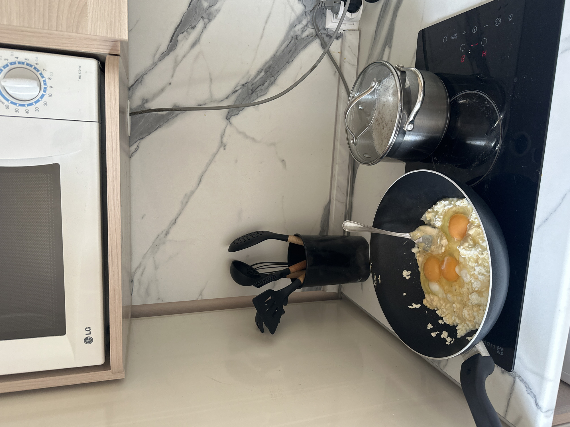 The image shows a kitchen scene from an elevated perspective. On the left, a white LG microwave is built into light wooden cabinetry. Adjacent to the cabinetry, a white countertop with gray marble veining extends towards the right. On this countertop, there is a black container holding cooking utensils. A black frying pan on a black stovetop contains cooking eggs with a fork resting in the pan. A stainless steel pot with a glass lid is placed on another burner of the stovetop. The stovetop control panel is visible with the number 14 displayed. The scene suggests cooking in progress in a domestic kitchen setting. The location and city name were not mentioned, therefore I can't include that information in this summary.