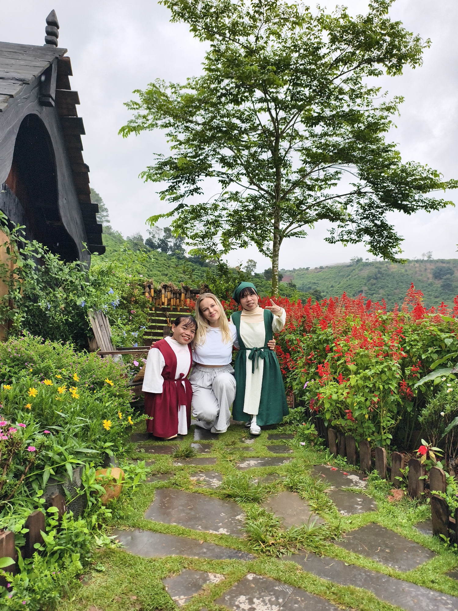 Three individuals pose on a stone path amidst a garden. One woman on the left wears a red and white garment. The woman in the center wears a white top and light gray pants. The woman on the right wears a green and white dress with a matching head covering, and she holds up her fingers in a "peace" sign. The background features a variety of flowering plants, with red flowers prominently displayed. A wooden structure is visible on the left, while a hillside with vegetation is in the background. The location is Thôn 1, Vietnam.