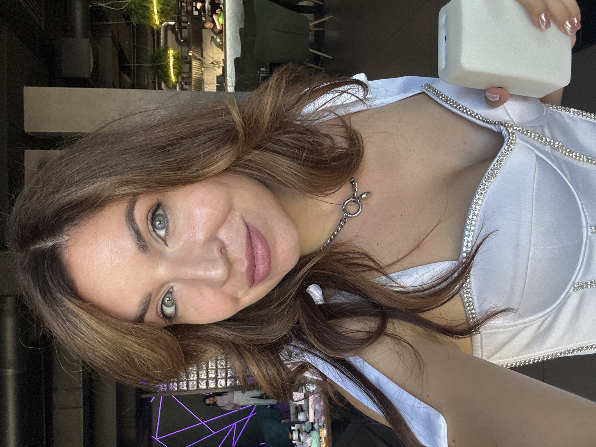 In Kryvyi Rih, Ukraine, a woman is captured in a close-up, selfie-style photograph. She has brown hair styled in loose waves and is wearing a white, low-cut top adorned with rhinestones. A silver necklace with a circular pendant rests on her collarbone. Her left hand is raised, holding a small, white, rectangular object. The background suggests an indoor setting, possibly a restaurant or cafe, with blurred figures and furniture visible. The lighting is bright, highlighting the woman's face and attire.