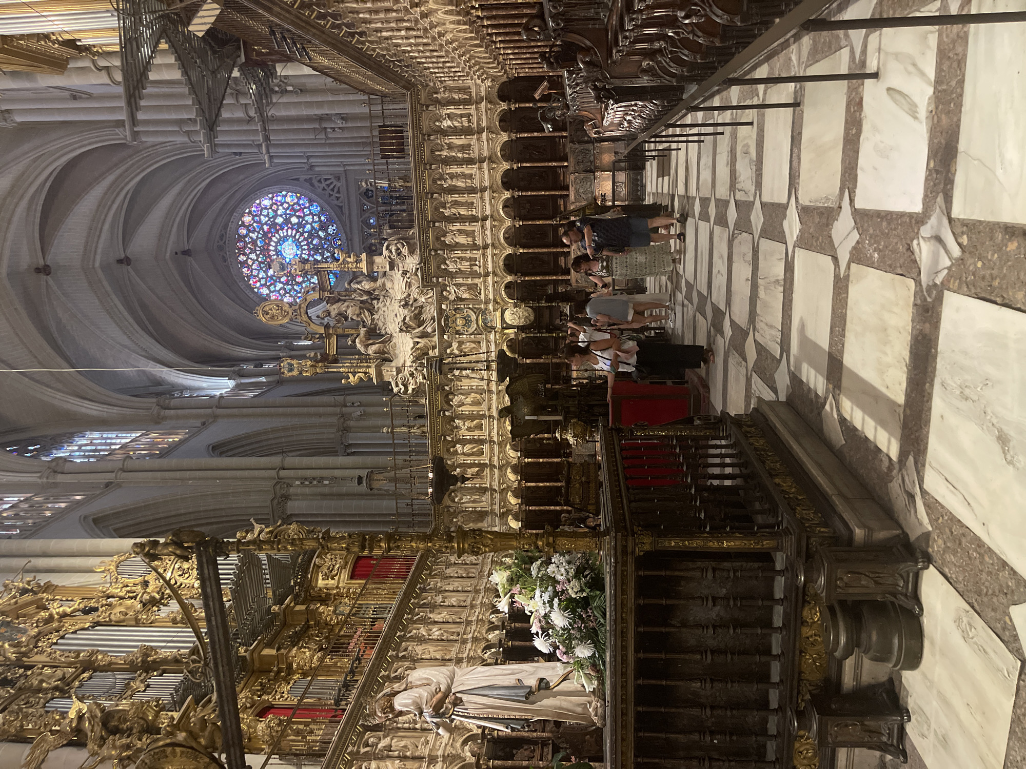 The image shows the interior of the Toledo Cathedral in Toledo, Spain, a Gothic cathedral renowned for its stunning architecture and rich history. The perspective is from within the nave, looking towards the altar and the rose window.
Dominating the left side of the image is a grand, ornate organ, crafted from gold and intricate metalwork, with red drapery visible behind it. In the foreground, to the left, stands a statue of a woman holding a child, draped in white fabric, with a bouquet of white and pink flowers placed at its base.
The central area of the cathedral is filled with elaborate choir stalls, featuring carved wooden seats and detailed sculptural panels depicting religious figures. Above the choir stalls, a large, colorful rose window, filled with stained glass in shades of blue, pink, and red, is visible in the distance, illuminating the soaring vaulted ceiling.
Several people are present in the scene, appearing to be tourists or visitors. They are scattered throughout the choir area, observing the architecture and artworks. One group of individuals is seen in the middle ground, while others are further back, closer to the altar. The floor is a checkerboard pattern of white marble tiles interspersed with darker stone. The overall impression is one of grandeur, historical significance, and a place of worship and art.