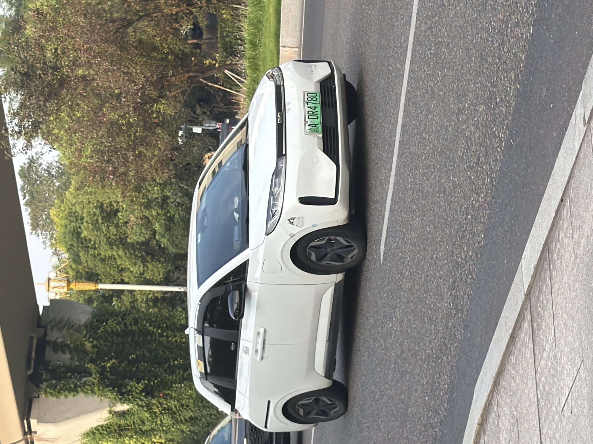 A white BYD hatchback is driving on a road in Jiangjin, China. The car has a license plate that reads "A DR4780". It is daytime, and the car is moving in the middle lane of a multi-lane road. The road has a sidewalk on the left side. There are trees and a bridge in the background. The car appears to be clean and in good condition. The overall scene is ordinary and shows a typical street view in an urban environment.