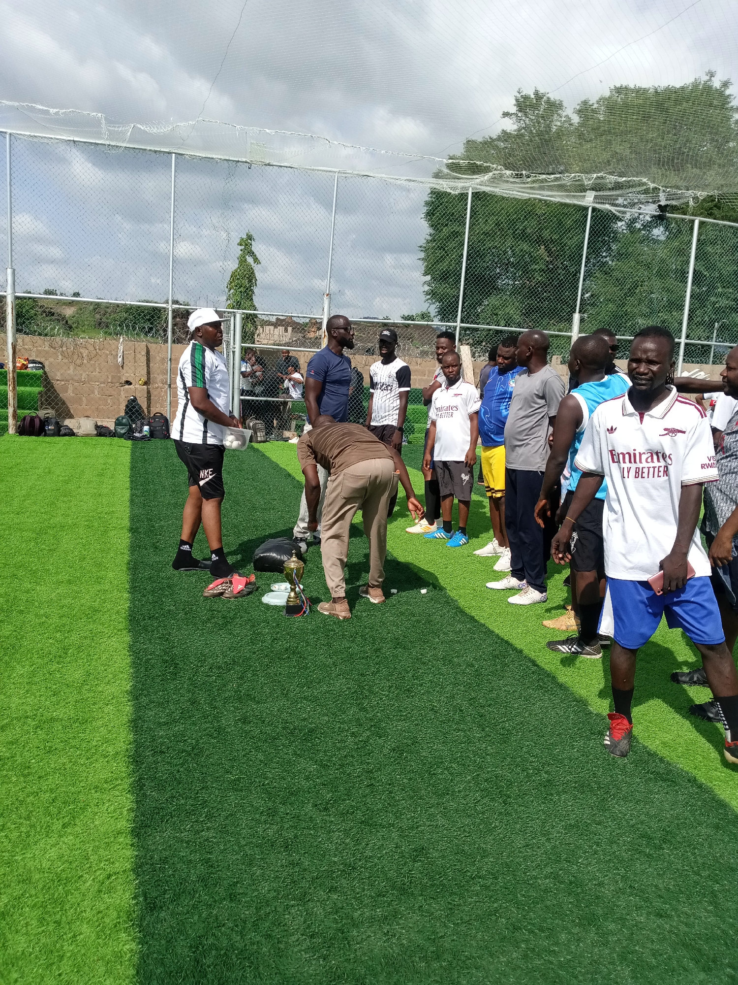 The image depicts a group of men gathered on an artificial turf field in Jimeta, Nigeria, possibly for a sporting event or a team meeting. The setting appears to be a fenced sports facility, with a net overhead and a brick wall in the background. A prominent trophy is placed on the ground in the foreground, suggesting an award ceremony or a competitive event has recently concluded or is about to commence. Several men are wearing athletic attire, including jerseys and shorts, and some have soccer cleats on. One man, wearing a white cap and a white and green shirt with black shorts, is holding a plastic container filled with white objects, possibly balls for a game. Another man in a white Emirates shirt and blue shorts is holding a pink mobile phone. The overall atmosphere seems to be informal and social, with individuals standing and conversing. The time of day appears to be daytime, with shadows cast on the field, indicating sunlight.
