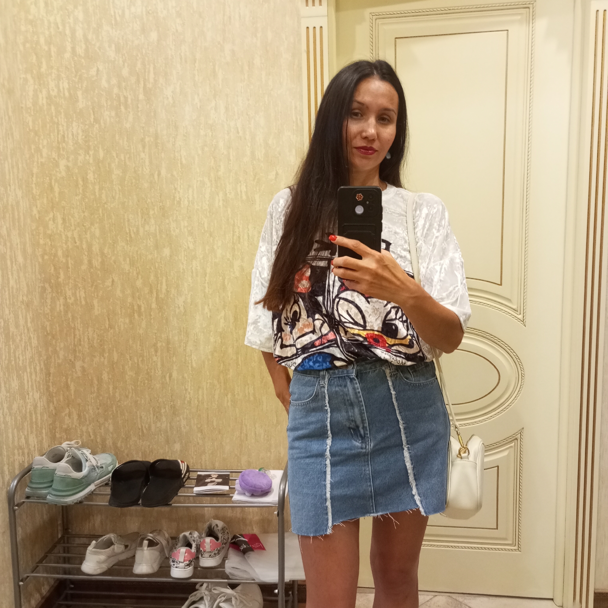 A woman is standing in front of a door and a shoe rack, taking a mirror selfie. She is wearing a white t-shirt with a cartoon graphic and a denim skirt. A white purse is slung over her shoulder. On the shoe rack are several pairs of shoes. The walls are a textured cream color, and the door is white with decorative molding. The scene appears to be an indoor entryway, possibly in Sochi, Russia.