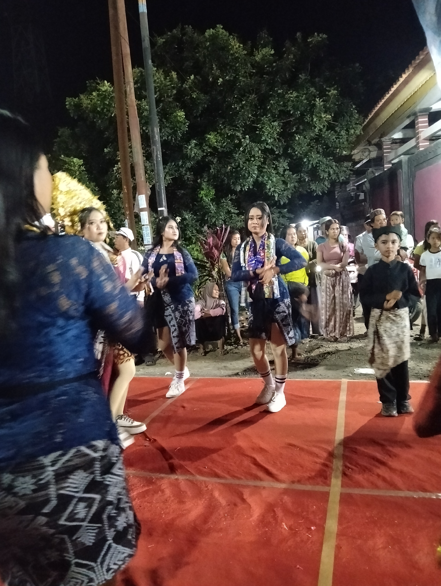 The image depicts an outdoor nighttime event in Sumbersewu, Indonesia. A group of people are gathered, with several individuals on a raised red platform, likely a stage. Three young women in matching dark blue and patterned outfits are performing a dance. They are wearing scarves around their necks and white athletic shoes with striped socks. One woman in the foreground on the left, also in a dark blue garment with intricate patterns, is partially visible and appears to be part of the performance or a participant. To the right of the dancers, a young boy in a black shirt and a patterned cloth around his waist is standing, facing forward. In the background, a crowd of onlookers is watching the performance, with some standing and others seated. Trees with lush green foliage are visible behind the stage, and a building with a reddish roof is to the right. Several poles, possibly for lighting or structures, are also present. The scene is illuminated by artificial lights, creating shadows on the red platform.