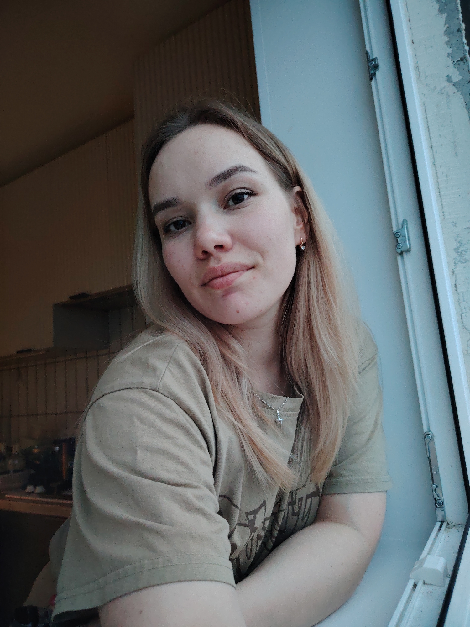 A young woman with light blonde hair and fair skin sits near a white window frame. She wears a beige t-shirt and a silver necklace with a star pendant. She has a small earring in her left ear. The background suggests an indoor setting, possibly a room with a cabinet or shelving unit visible. The location is Марковский, Russia.
