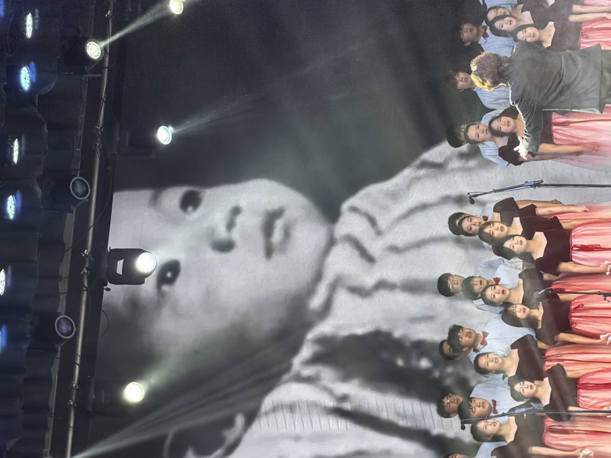 This image depicts an indoor musical performance taking place on a stage in Guta, China. The scene is dominated by a dramatic interplay of light and shadow.
On the right side of the frame, numerous performers, likely a choir or vocal group, are arranged in multiple rows. The female performers wear dark tops paired with long, flowing pink or reddish-pink skirts, while the male performers are dressed in light-colored shirts with red bow ties. In the upper right, a male conductor in a dark suit jacket, with his back mostly to the viewer, is actively directing the ensemble with an uplifted arm. Several stand microphones are visible in the foreground, positioned in front of the performers.
On the left side, a large black and white image of a person's face is projected onto a screen or backdrop. The face, shown in a three-quarter view, carries a serious or contemplative expression. Bright stage lights, some emitting white/yellow beams and others blue light, are mounted on the far left, casting distinct rays across the stage and illuminating parts of the projected image. The overall lighting is theatrical, with the stage and performers brightly lit against a darker background. The image captures a focused and formal performance, possibly a concert or a tribute event. No visible text is present.