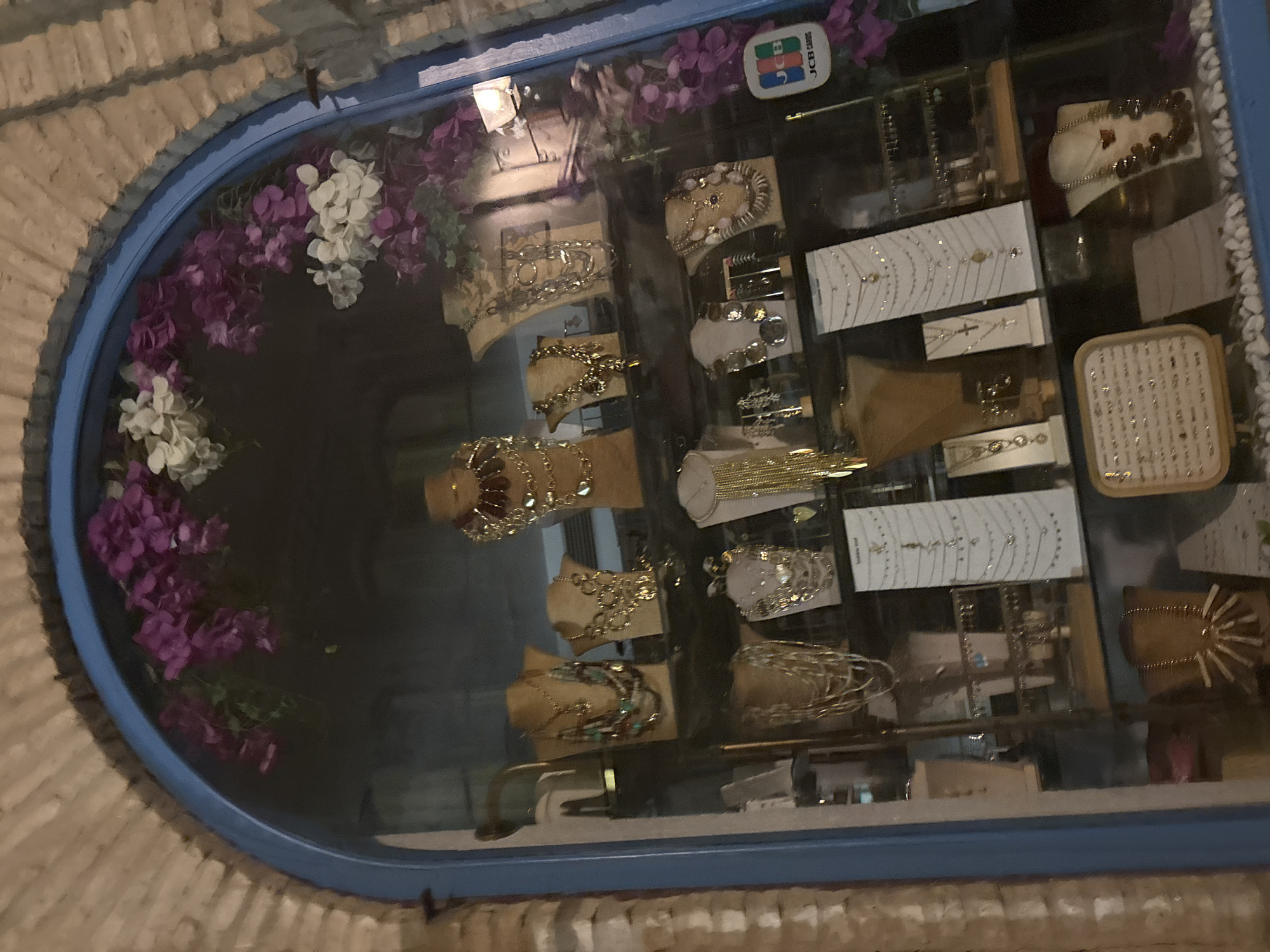 A jewelry display case is seen through a blue archway, likely in the city of Córdoba, Spain, as suggested by architectural details. The case holds various jewelry pieces, including necklaces, bracelets, and possibly earrings, presented on stands or cards within the glass display. The case is framed with artificial white and purple flowers, and is illuminated by a light source. A sticker with "JCB Cards" is visible at the upper edge of the display case. The surrounding structure appears to be constructed from light-colored brick.