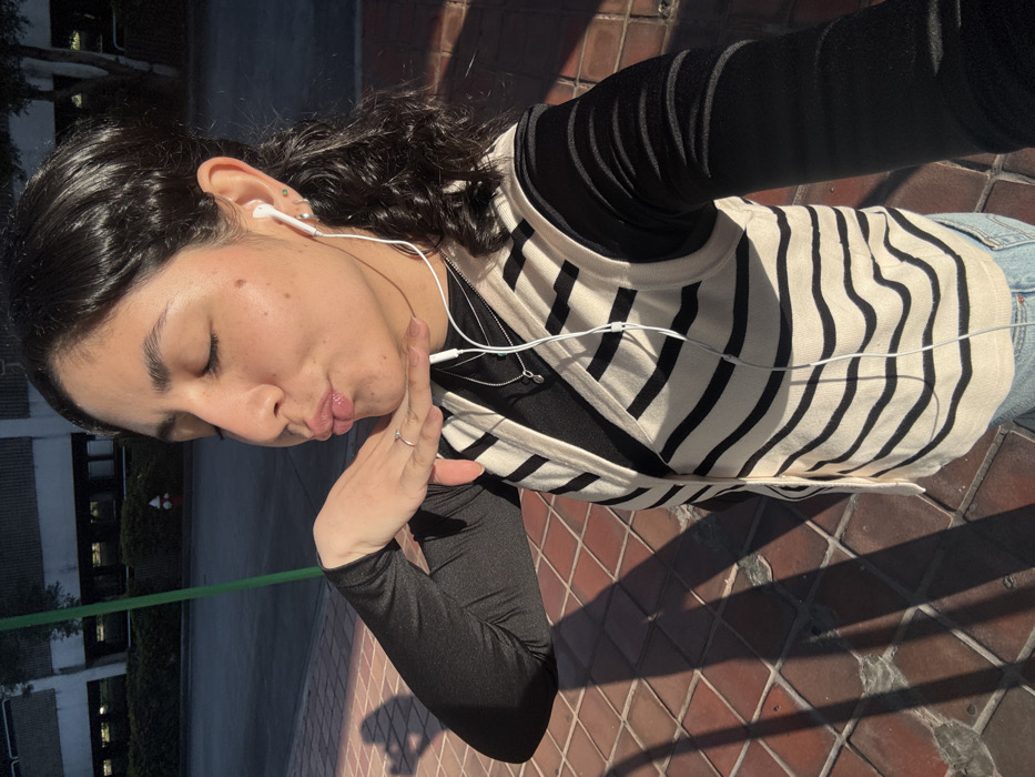 The image displays a selfie-style portrait of a young adult female, positioned outdoors under direct sunlight. The subject occupies the majority of the frame, angled slightly to her right, with her eyes closed and lips puckered in a "kissing" pose. Her right hand is positioned below her chin. She wears a black long-sleeved shirt underneath a cream-colored, V-neck vest with horizontal black stripes, and light blue jeans are visible at the bottom of the frame. White wired earbuds are inserted into both ears, with the wire extending down her chest over a thin silver-colored necklace featuring a small circular pendant. A silver-colored ring is present on her right ring finger.
The foreground consists of a reddish-brown brick or tiled sidewalk, where long, distinct shadows from the subject and a green pole are cast by direct sunlight from the left. Beyond the sidewalk, an asphalt road is visible. In the background, a multi-story building with a light-colored facade and numerous windows is partially obscured by dark green foliage at ground level. A red fire hydrant is visible on the right side of the road in the background. The scene is located in the city of Hialeah, United States of America.