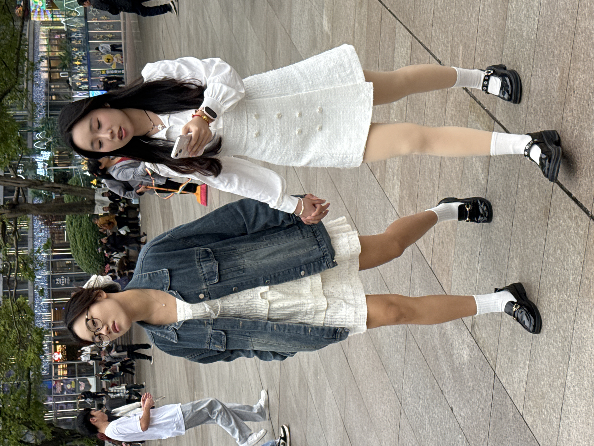 Two young women are standing on a tiled pedestrian walkway in what appears to be an urban environment. The woman on the left is wearing a white dress, a denim jacket, white socks, and black shoes. She has dark hair pulled back in a ponytail and wears glasses. The woman on the right is wearing a white dress with button details, a long-sleeved white shirt, white socks, and black shoes. She has long, dark hair and is looking at a smartphone in her hand. They appear to be holding hands. In the background, there is a building with neon lights and a crowd of people. The presence of the Chinese writing suggests this could be a city in China. Based on the appearance of the background architecture and neon signage, the location could be the Liangjiang New Area in Chongqing, China. There are other people walking around in the background, and trees are visible on the left.