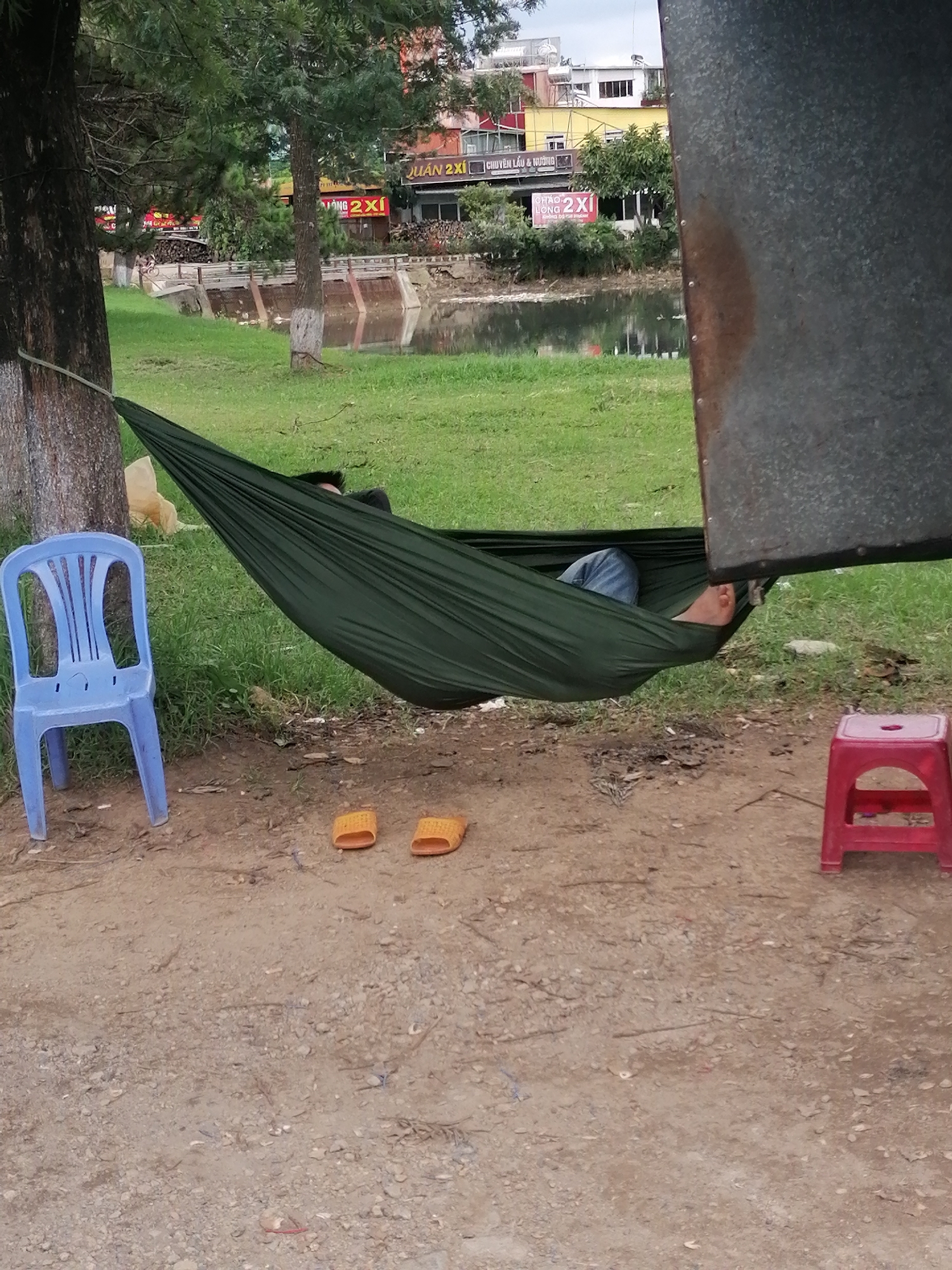 A person is lying in a green hammock stretched between two trees. The hammock is suspended over a dirt and gravel area. To the left of the hammock is a bright blue plastic chair. Two orange flip-flops are placed on the ground in front of the hammock. To the right of the hammock is a small red plastic stool. In the background, there is a body of water with a concrete embankment. Across the water, there are buildings with red roofs and signs written in Vietnamese. The scene appears to be in Da Lat, Vietnam, with the characteristic greenery and architecture. A large, dark, metallic object partially obscures the right side of the image.