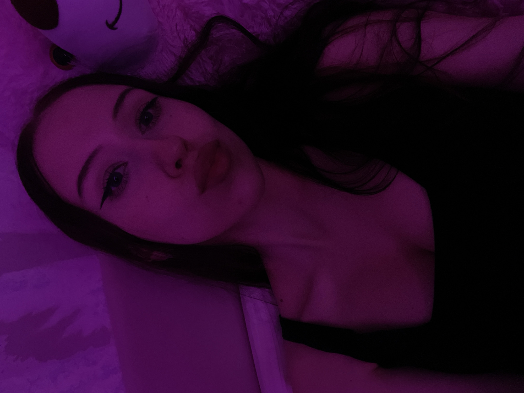A young woman with long, dark hair is lying down, facing the camera. She is wearing a black top. Her face is illuminated by a purple light, and she is making a kissing gesture with her lips. Behind her, a plush toy, possibly a bear, is visible. The background appears to be a textured surface with a purple hue. The image is a selfie, taken in a dimly lit room. There is no discernible information about the location being Votkinsk, Russia.
