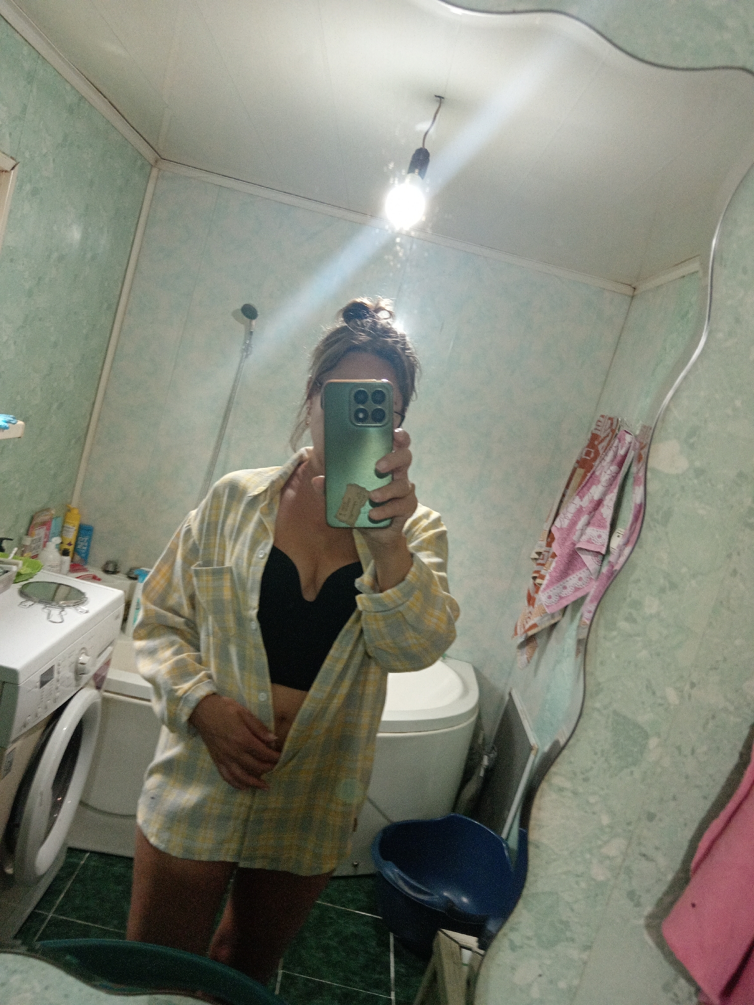 A woman is taking a selfie in a bathroom. She is wearing a yellow and white plaid shirt unbuttoned over a black bra and is holding a green smartphone in her right hand. The bathroom has light green tiled walls and white ceiling. A light bulb hangs from the ceiling. To the left of the woman, a washing machine is visible. To her right, a large, warped mirror reflects the scene. A toilet and a blue plastic basin are also present in the bathroom. The overall impression is of a domestic, casual setting.