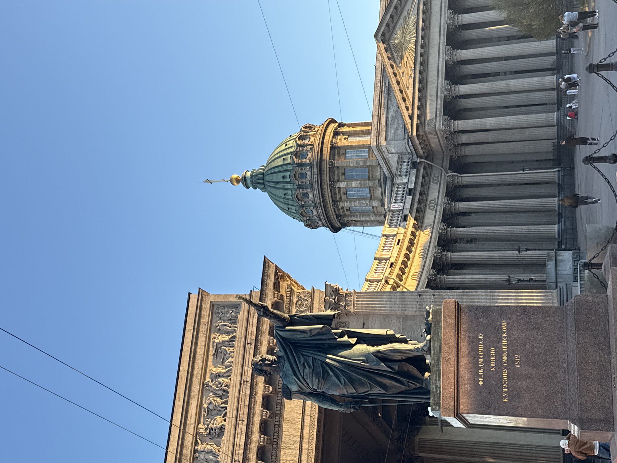The image shows the Kazan Cathedral in Saint Petersburg, Russia. In the foreground is a statue of Mikhail Kutuzov, a field marshal in the Russian army. The cathedral's dome is visible, as are some of its columns and architectural details. A few people are seen walking along the sidewalk next to the cathedral. The sky is clear and blue.