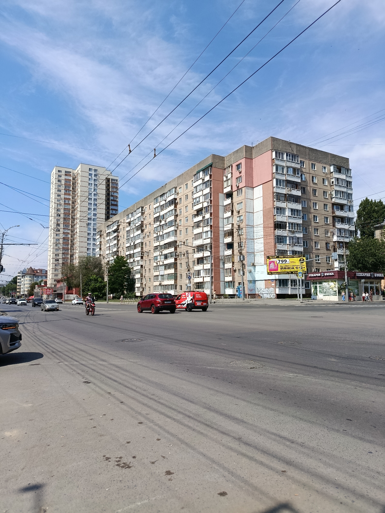 The image depicts a street scene in Saratov, Russia, on a sunny day with a blue sky and scattered clouds. The foreground shows a wide asphalt road with tire marks and some oil stains. In the middle ground, there are several multi-story apartment buildings, one of which is particularly tall and slender, while another is a long, block-style building with pink and grey facades. Various vehicles are visible on the road, including a red SUV, a motorcycle with a rider, a silver car, and a red van. Several people are walking on the sidewalks near shops. Overhead, power lines crisscross the sky, casting shadows on the road. One building has a large yellow advertisement banner with Cyrillic text and prices. Another building has storefronts with signage indicating a bakery. The environment suggests an urban residential and commercial area.