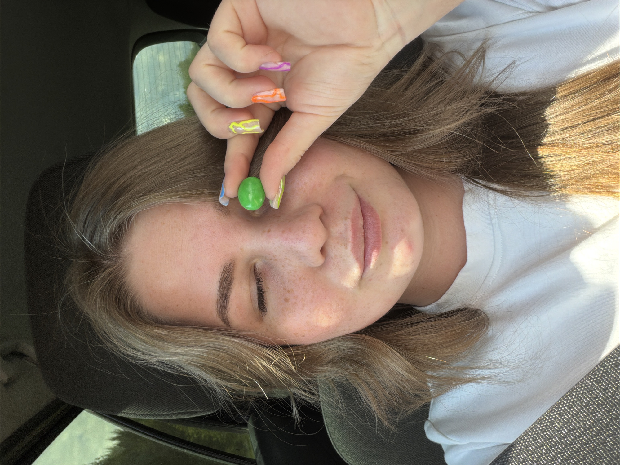 A young woman with blonde hair and freckles is sitting in the driver's seat of a car. She is holding a green candy between her thumb and index finger, bringing it up to her left eye. Her left eye is closed, and she is smiling slightly. Her nails are painted with bright, colorful patterns. The background shows the interior of the car and a glimpse of the outdoors through the windows, suggesting a rural or wooded setting. The location is identified as Oreshniki, Russia.