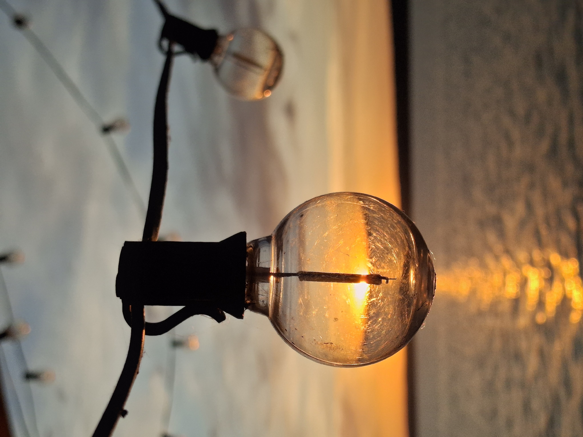 The image features a string of decorative light bulbs, with the primary bulb in sharp focus. The filament of the bulb is visible, reflecting a bright orange light, likely from the setting sun, creating a warm glow within the glass. This light also casts a diffused, shimmering reflection on a textured surface in the background, possibly water or a textured wall. Another bulb, slightly out of focus, is visible behind the main one, as well as additional string lights in the distance. The setting suggests an outdoor location in Chaikovsky, Russia, at dusk or dawn, given the quality and color of the light. There are no people in the frame.