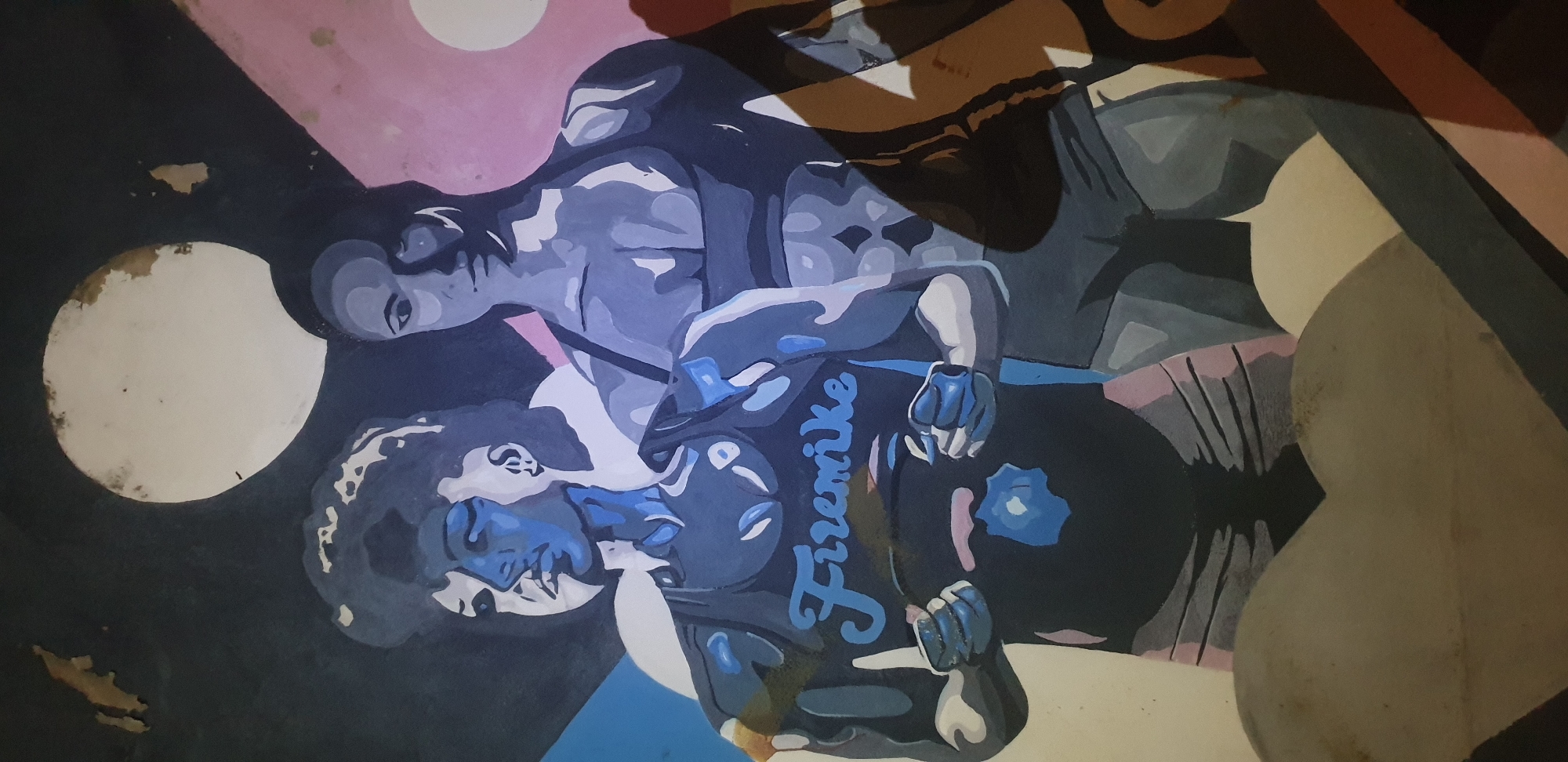 The image displays a mural painting in Narayi, Nigeria, featuring two individuals rendered in shades of blue and gray. The person on the left, with dark curly hair, wears a black tank top with the word "Firenike" in blue lettering. This individual's skin tone is depicted in deep blue and lighter blue highlights. The person on the right, with short dark hair, is shown in a gray sports bra and dark shorts. Their skin is primarily gray with darker gray shading. Both individuals appear to be standing. The background is abstract, with geometric shapes in shades of black, pink, and blue, and a large white circle resembling a moon or sun. There are signs of wear and tear on the mural, with peeling paint visible in the upper left section.