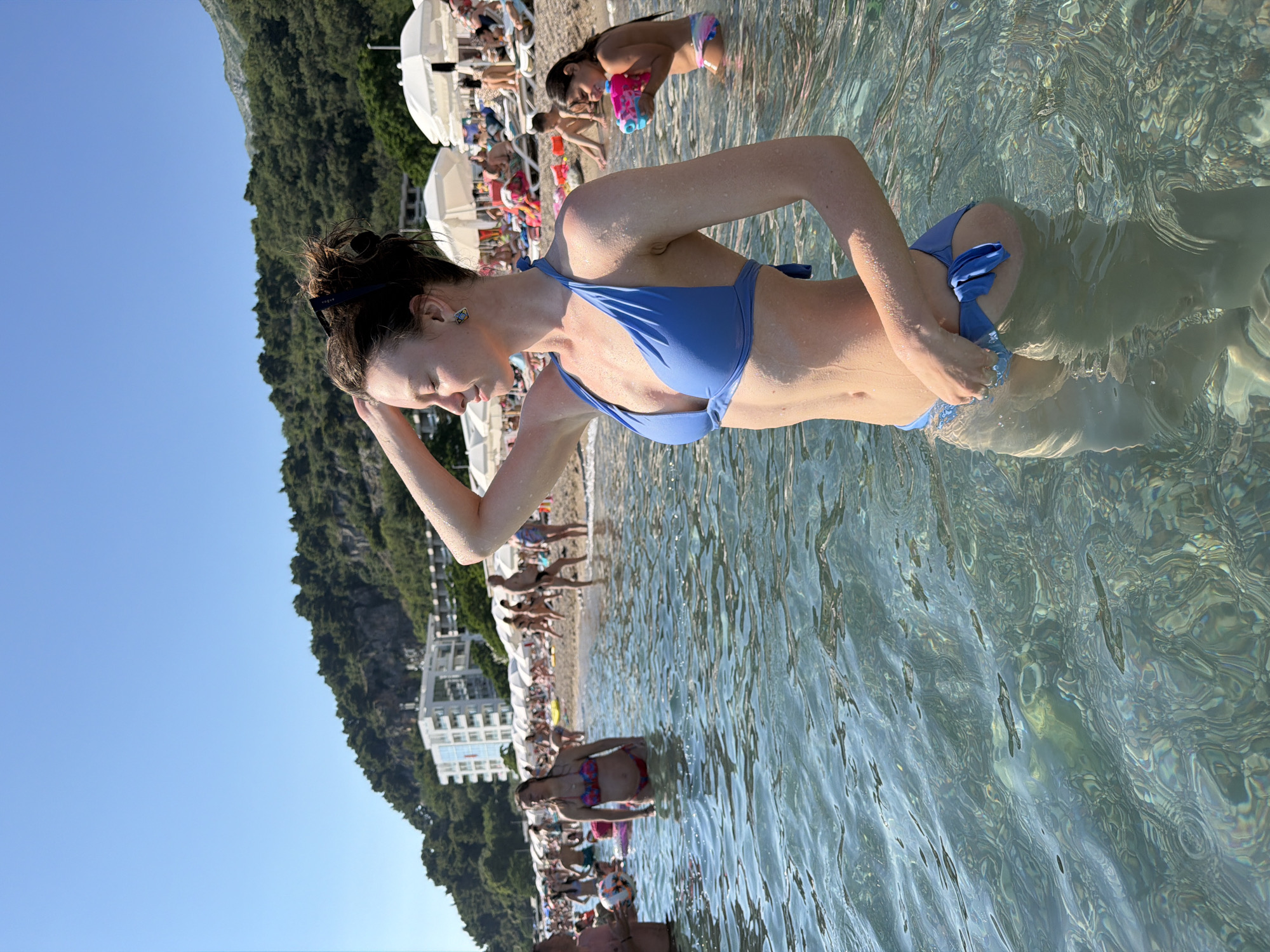In Sutomore, Montenegro, a young woman stands waist-deep in the clear Adriatic Sea, facing right with her eyes closed. She wears a blue bikini and has her left arm raised, resting her hand on her head. The water is calm, revealing the pebble-strewn seabed below. In the background, a crowded beach stretches along the coast, with numerous beach umbrellas, lounge chairs, and people relaxing under the bright sun. A multi-story white hotel is visible further up the shoreline, backed by a steep, tree-covered hill. The sky is a clear, vibrant blue, indicating fair weather.