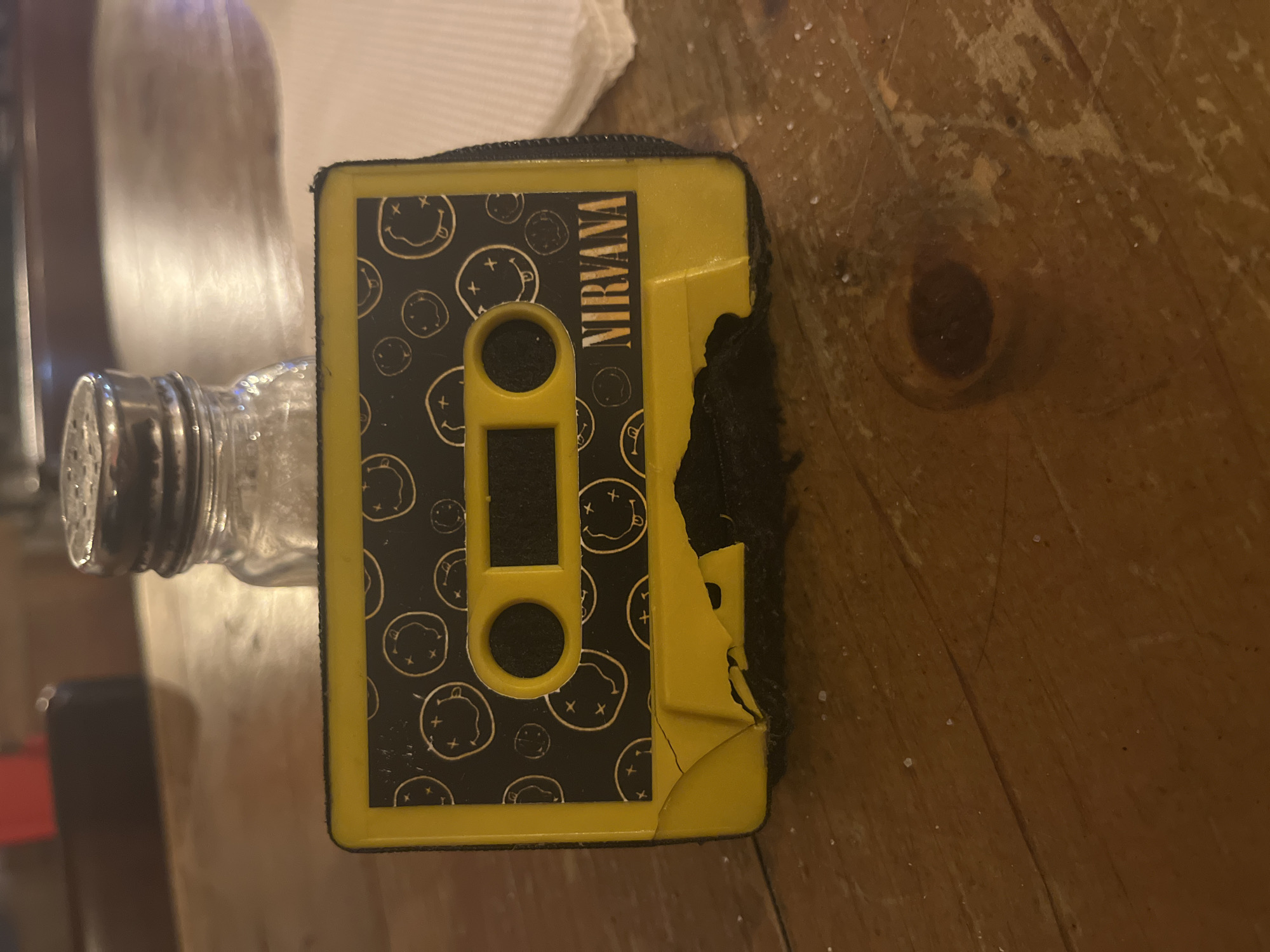 The image shows a yellow and black cassette tape case with the word "NIRVANA" printed on the side. The case is patterned with smiley faces that have crossed-out eyes. The case is slightly damaged on the right side, with the yellow plastic peeling away. The case is sitting on a wooden surface that appears to be a table. A glass salt shaker is partially visible to the left of the cassette case. The surface of the table has a few white specks on it. The image does not provide any information to determine the location in Barcelona, Spain.