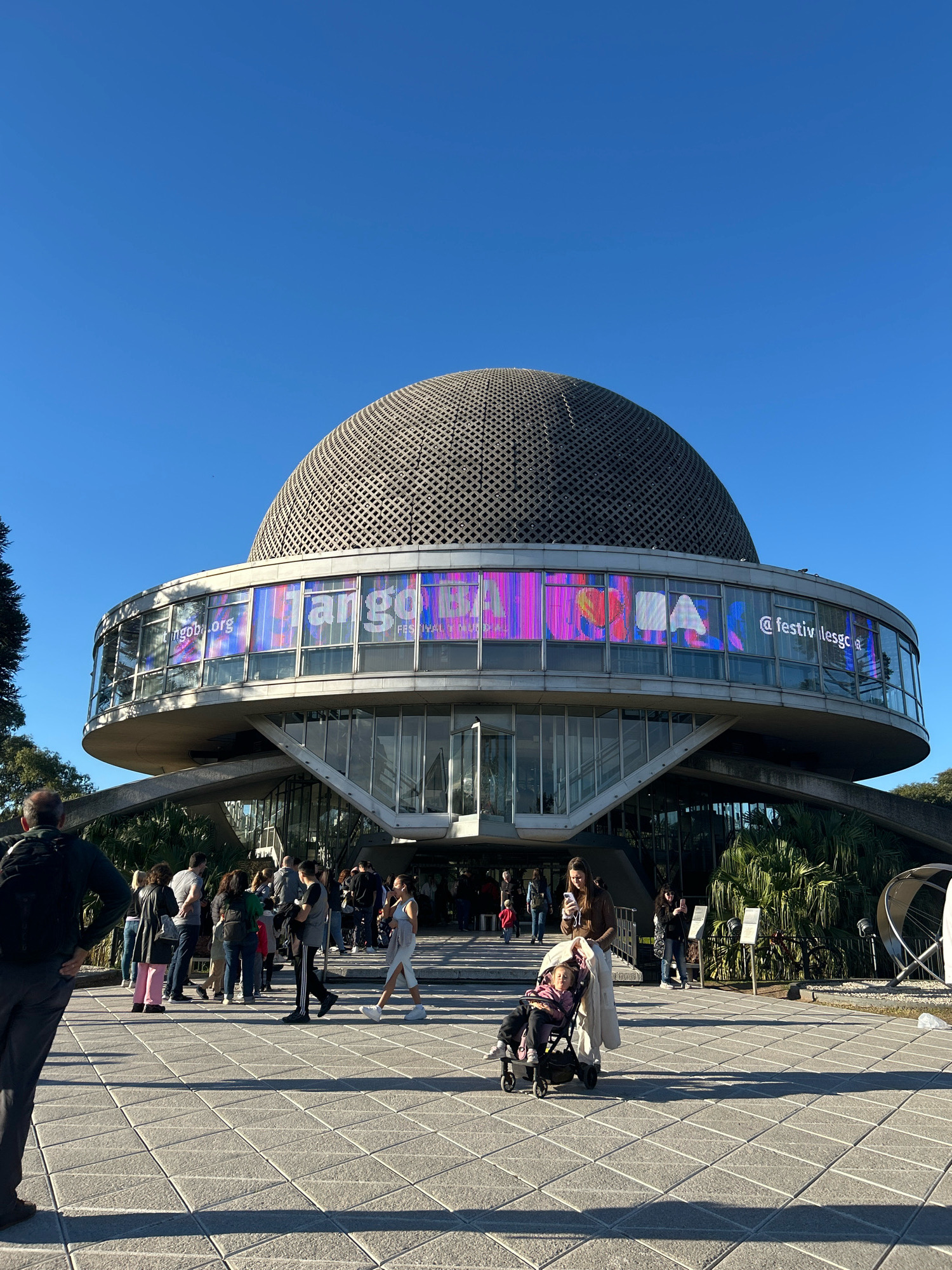 The image shows the Planetario Galileo Galilei, located in the Palermo neighborhood of Buenos Aires, Argentina. A large crowd of people is gathered in front of the building, which features a prominent dome and a circular structure with large windows displaying a vibrant digital advertisement. The sky is clear and blue, and the ground is paved with hexagonal tiles. Several individuals are visible, including a woman pushing a baby stroller, a man with a backpack, and a group of people talking and walking. The building's architecture is modernist, with concrete and glass elements. Lush green vegetation surrounds the area, and shadows are cast across the tiled plaza, indicating a sunny day.