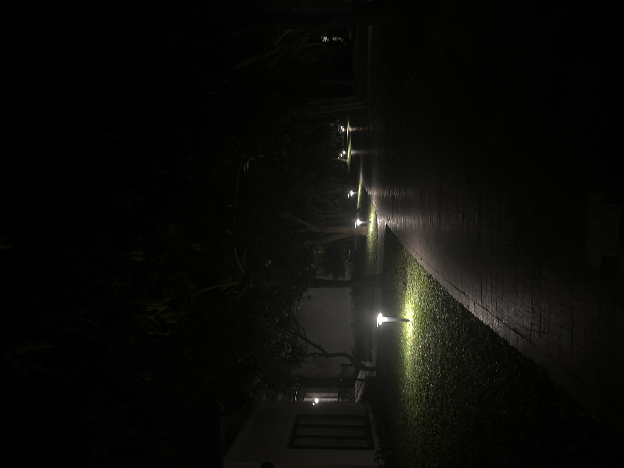 The image depicts an outdoor scene at night. A dimly lit brick pathway stretches into the darkness, lined on one side by grass illuminated by a series of low-level lights. Trees and foliage are visible on the left side of the path, merging into the surrounding darkness. In the foreground, a building with a visible window is partially illuminated by the nearest light. The location appears to be a residential area, possibly in the city of Unknown, Unknown. The environment is dark and quiet, with the only visible activity being the illumination of the path and surrounding vegetation.