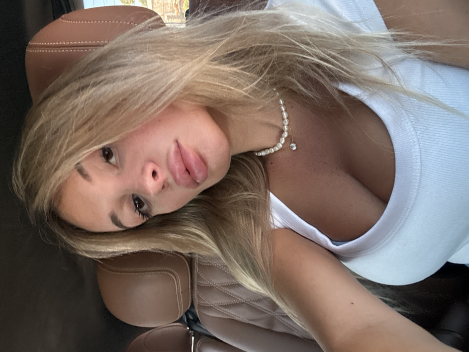 A young woman with long blonde hair is reclining in what appears to be a car seat. She is wearing a white tank top and a pearl necklace with a small diamond pendant. Her head is tilted towards the camera, emphasizing her full lips and dark eyes. The background shows the light-colored leather of the car seat, and the overall lighting is bright and airy.