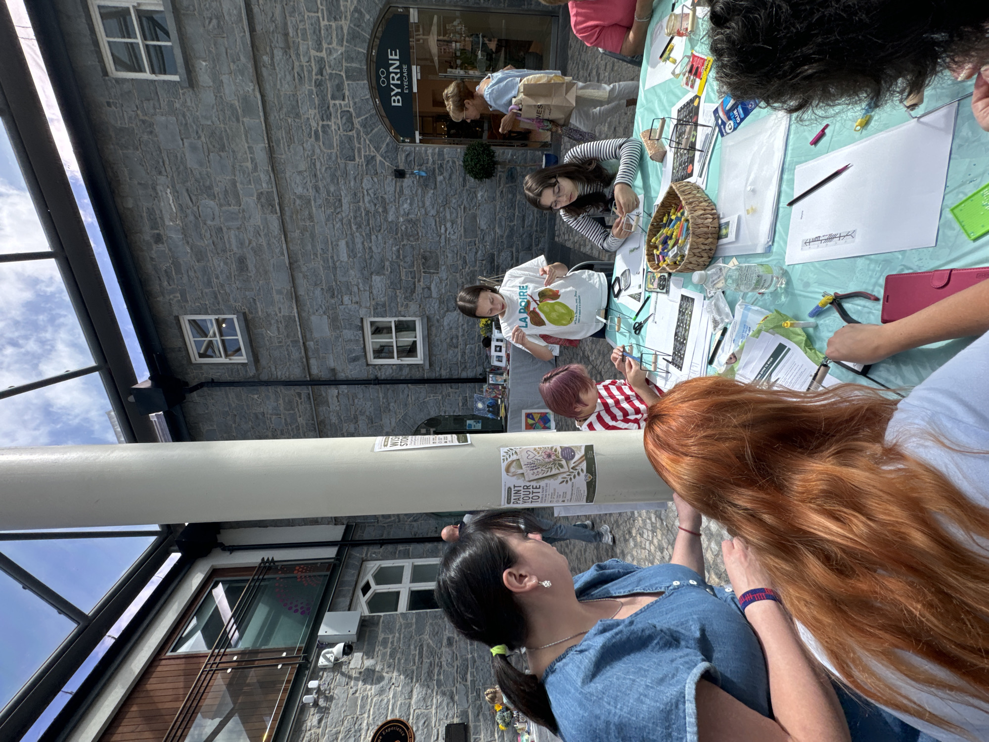 The image depicts an outdoor craft event taking place in Kilkenny, Ireland, likely at the Kilkenny Design Centre. Several people, including adults and children, are gathered around a table covered with a teal-colored cloth. They appear to be engaged in crafting activities, with various art supplies, tools, and materials spread across the table, such as beads, pliers, and paper. In the background, the distinctive stone architecture of Kilkenny is visible, along with signage for "BYRNE EYECARE." The event is set under a glass-covered roof, suggesting it's an open-air atrium or courtyard. The atmosphere appears to be relaxed and participatory, with individuals focused on their creative endeavors.