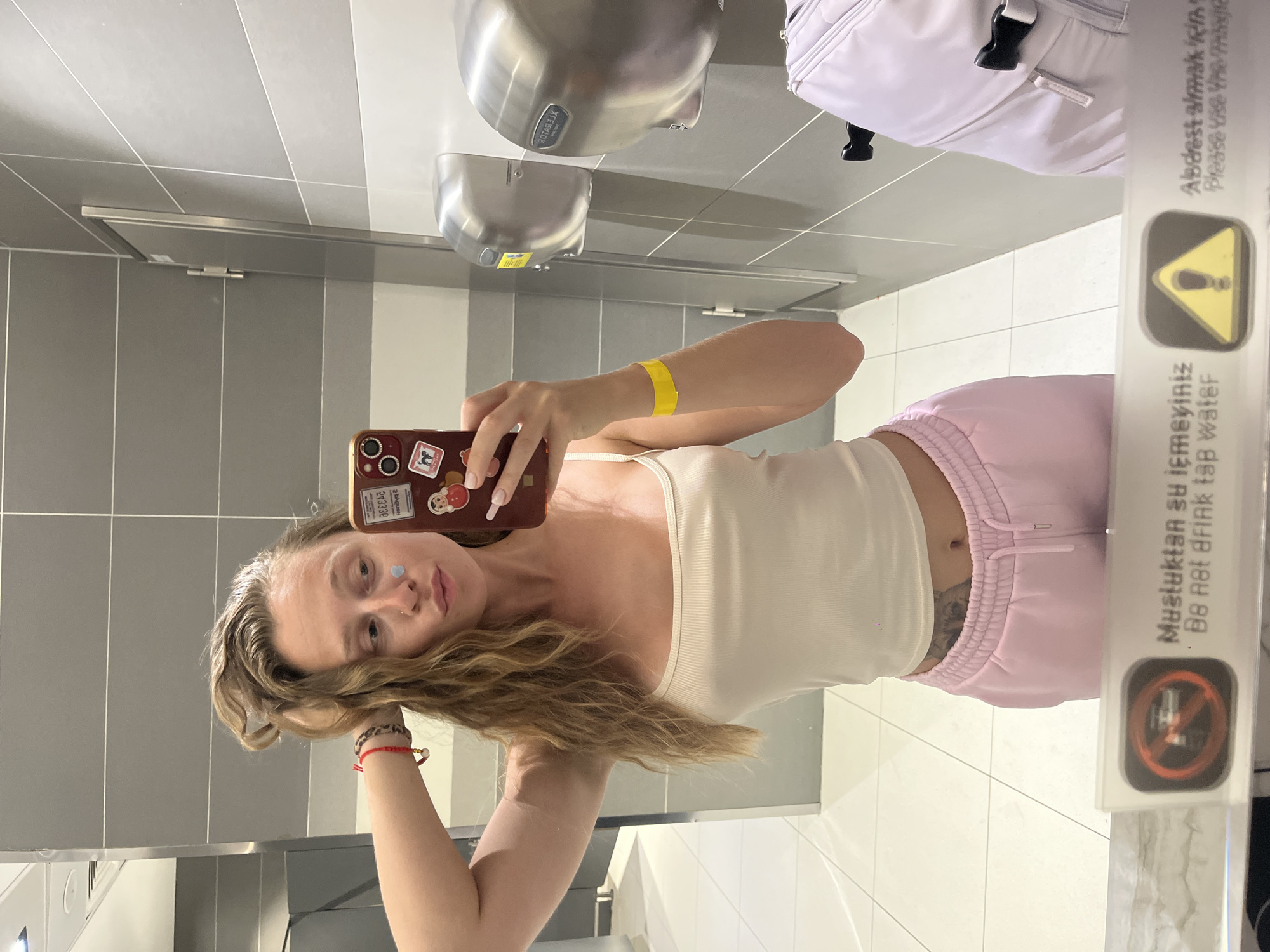 In a clean, modern restroom with grey tiled walls and white tiled floors, a young woman is taking a selfie in a mirror. She has long, wavy blonde hair and is wearing a light-colored ribbed tank top and pale pink sweatpants. She is holding her left hand up to her hair and her right hand is holding a red-cased smartphone, which is angled to capture the reflection. On her face, there is a small blue sticker or cosmetic applied to her left cheekbone. Around her left wrist, she wears a red string bracelet with beads, and around her right wrist, a yellow band. The background of the restroom includes a hand dryer mounted on the wall. A sign in the foreground, visible in the reflection, shows a prohibition symbol with a drink and text in Turkish and English reading "Do Not Drink Tap Water" and a yellow triangle with an exclamation mark, also with text in Turkish and English indicating the need to use the provided water. To the right, a portion of a white piece of luggage is visible. The overall impression is that of a traveler taking a moment to document herself in a public restroom, likely at an airport or similar transit hub in Arnavutköy, Turkey, given the context of the sign's text and the presence of luggage.