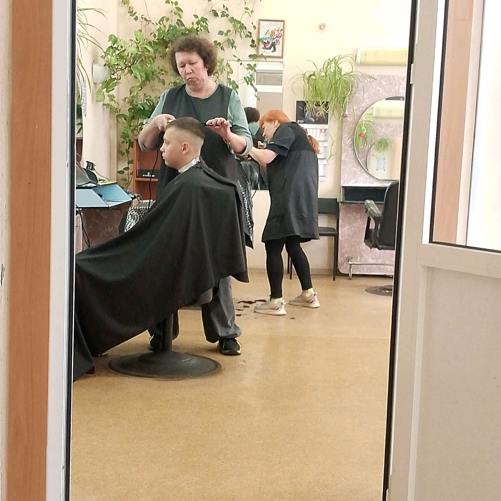 The image depicts a reflection of a hair salon in Vyazniki, Russia. Two women are visible, one adult with curly brown hair, wearing a dark blue apron over a lighter blue top, is actively cutting the hair of a young boy. The boy, facing left, is seated and covered by a black cape. He has short, neat hair that is in the process of being styled.
A second woman with red hair, also wearing a dark apron, is partially visible in the background, engaged in a similar hair cutting activity. The salon environment is suggested by the presence of styling chairs (one with a black seat and metal base is prominent), shelves with items, and plants. A round mirror is visible on the back wall. The floor appears to be tiled or linoleum, with scattered hair clippings around the boy's feet. The reflection is captured through an open doorway, with the frame of the doorway visible in the foreground.