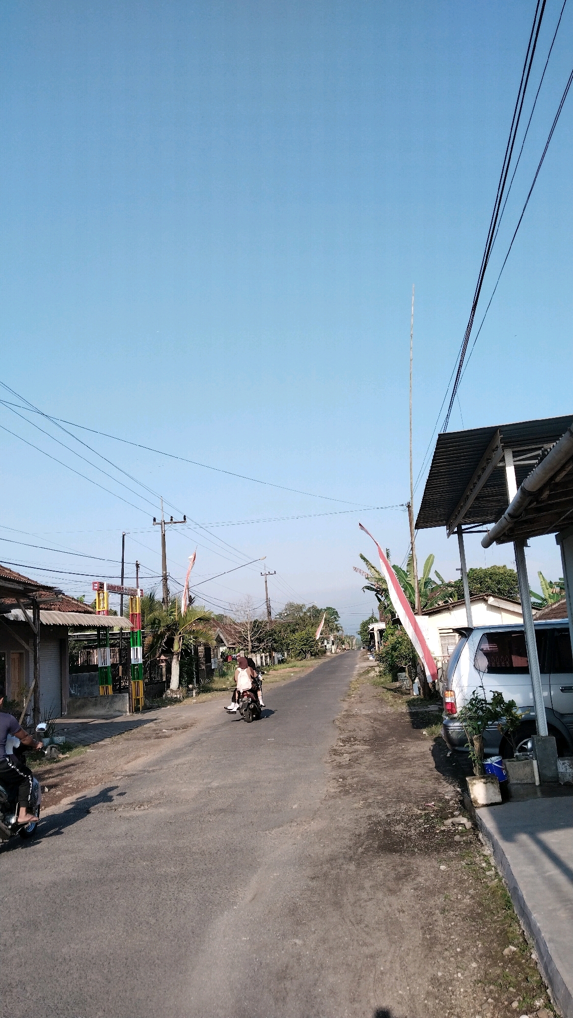 The image shows a street scene in Kaligondo, Indonesia. A paved road runs through a village with houses and trees on either side. The sky is clear and blue. Two people are riding motorcycles on the road, and a car is parked on the right side of the road. There are also power lines and utility poles visible throughout the scene. The overall impression is a peaceful, rural setting.