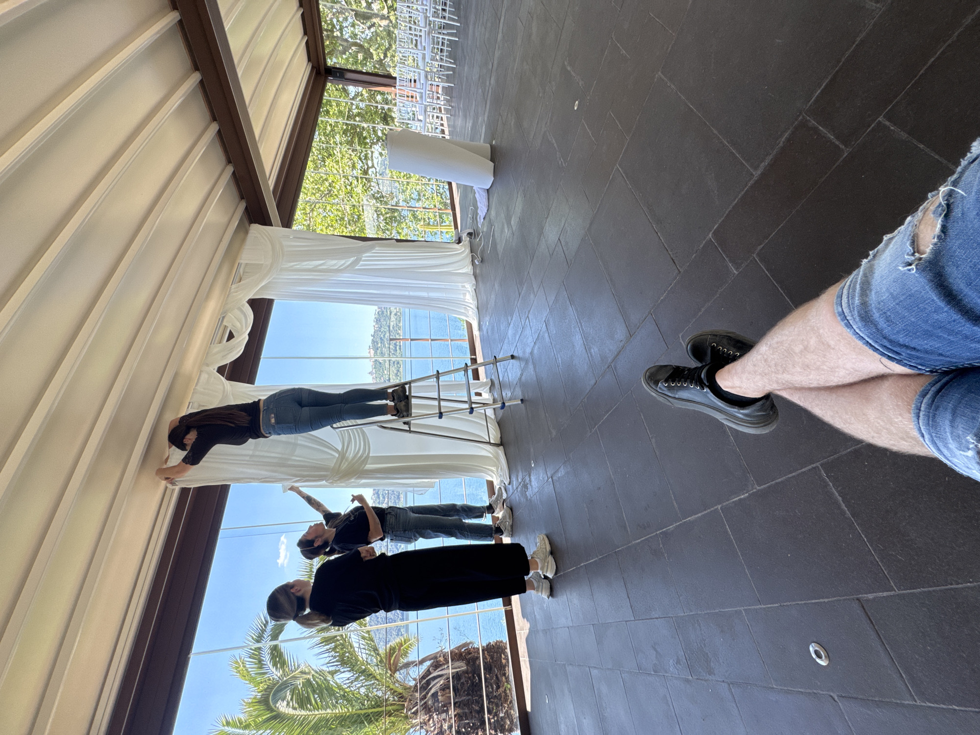 From an oblique angle, an interior view suggests a location in Castel Gandolfo, Italy. Three individuals are present. One is standing on a ladder, likely adjusting white curtains in front of large windows that overlook water and a tree-lined horizon. Another person is standing below on the ground, possibly assisting. A third figure is partially visible. The location seems to be a venue prepared for an event, based on the chairs and decorations visible. The flooring consists of dark, rectangular tiles. At the far right of the frame are the legs of a person wearing denim shorts and black shoes, who appears to be observing the scene. A large palm tree is seen in the foreground outside of the glass windows.