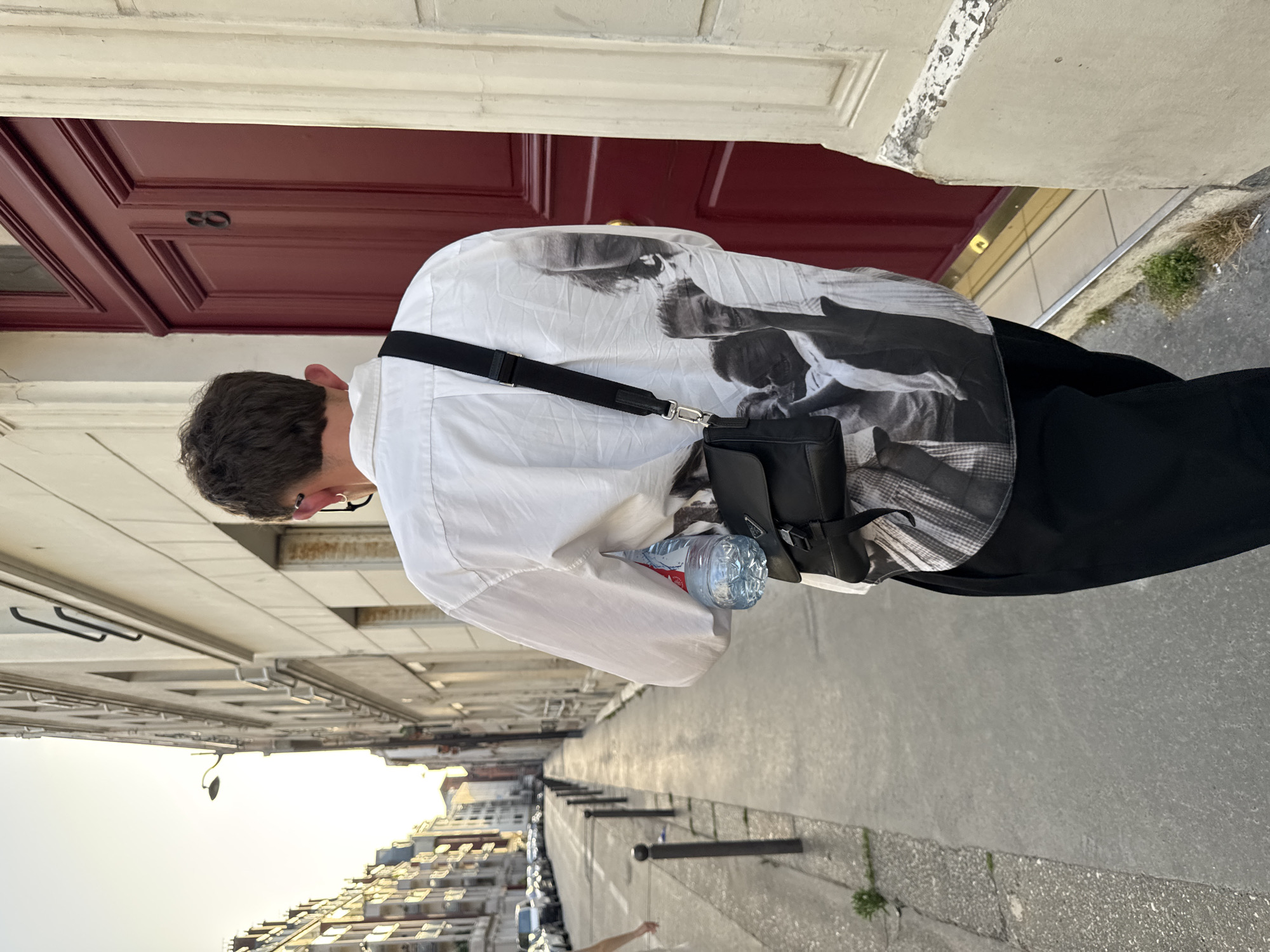 A person wearing glasses, a white shirt with a black and white print on the back, and black pants is standing on a sidewalk next to a street. The person has a black shoulder bag and is holding a water bottle. The sidewalk is lined with metal posts and greenery. In the background, there are buildings and parked cars. The image appears to have been taken on a street in Paris, France, as indicated by the architectural style of the buildings and the overall urban environment. A maroon door with the number "8" is also visible in the background.