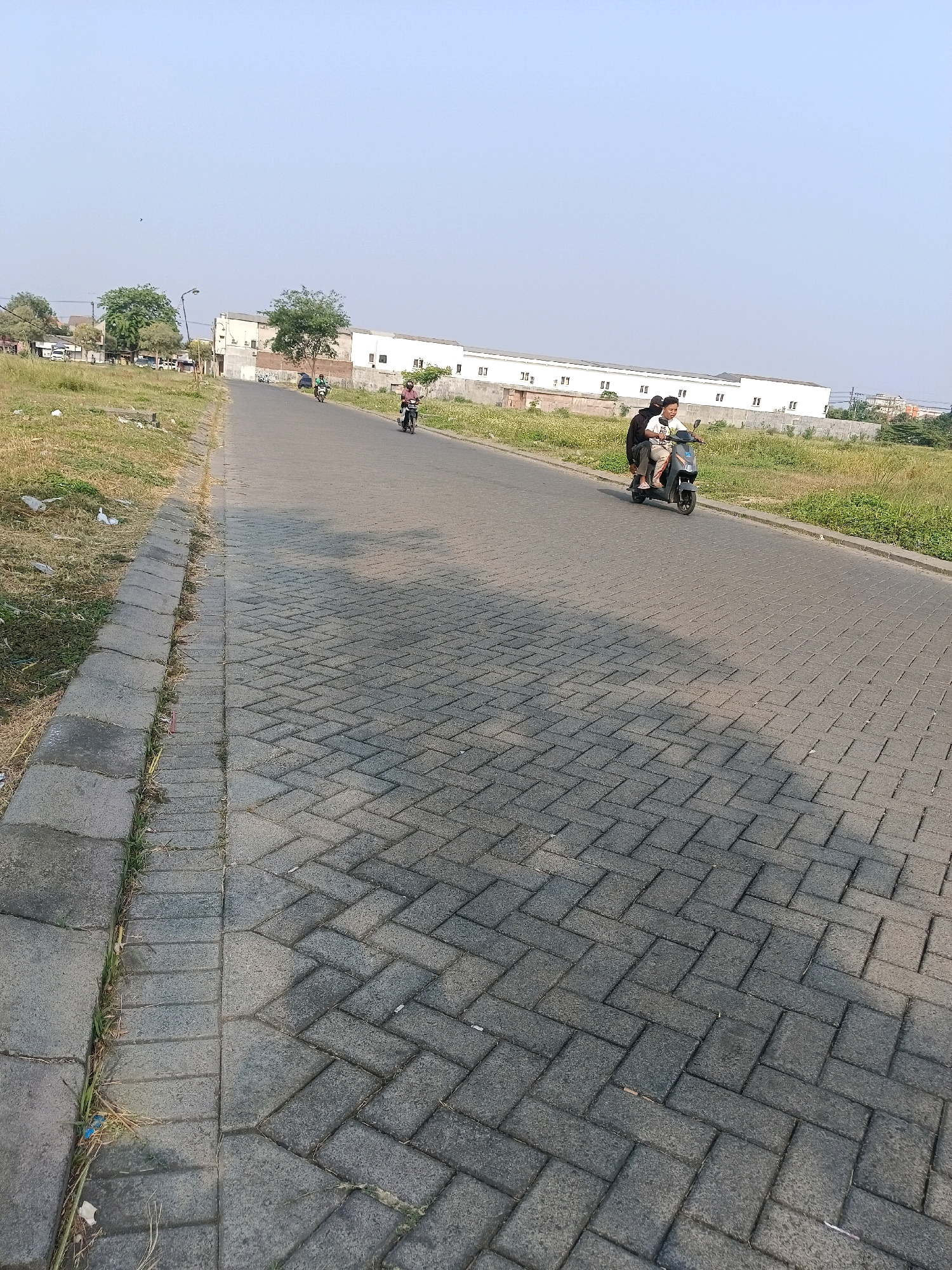 In Tawangsari, Indonesia, on a paved road with a brick-patterned sidewalk, two individuals ride a scooter. The scooter, dark grey, is being ridden by a young man wearing a white t-shirt and a black backpack. Behind him, a second individual, also appearing to be a young man, is seated and wearing a black hoodie. They are traveling away from the camera on the right side of the road. The road is flanked by a grassy area with some dry vegetation and scattered litter. In the background, several buildings with white facades and a few trees are visible under a clear, light blue sky. Another scooter with a single rider is further up the road on the left.