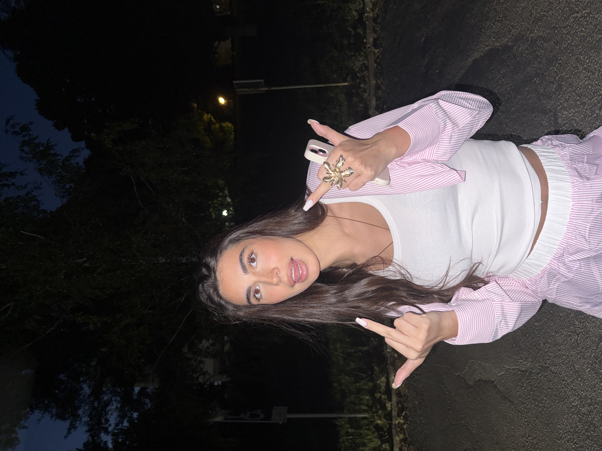 A young adult female, approximately 20-30 years old, stands outdoors at night in Blagoevgrad, Bulgaria, looking directly at the camera. She has long, dark brown hair parted down the middle, and prominent dark eyebrows. Her eyes are wide open, and she is sticking her tongue out slightly. She wears a white ribbed crop tank top, a light pink and white striped long-sleeved button-up shirt worn open over it, and matching striped shorts or pants with an elastic waistband. A small silver navel piercing is visible.
Her left hand is extended forward, displaying a middle finger gesture, with long, light-colored polished nails. Her right hand holds a light-colored smartphone (possibly an iPhone) with her index and pinky fingers extended, while her thumb, middle, and ring fingers are curled. A large, ornate, gold-toned floral or starburst-shaped ring adorns her right index finger.
The background is dark, featuring silhouettes of trees against a very dark blue night sky. A single illuminated orange/yellow streetlight is visible in the mid-ground on the right. Faint, indistinct lights are present in the distance. Two dark, vertical poles, possibly utility poles or signs, are visible in the background behind her. The ground is a dark asphalt surface. The scene is illuminated by artificial light sources, casting some highlights on the subject.