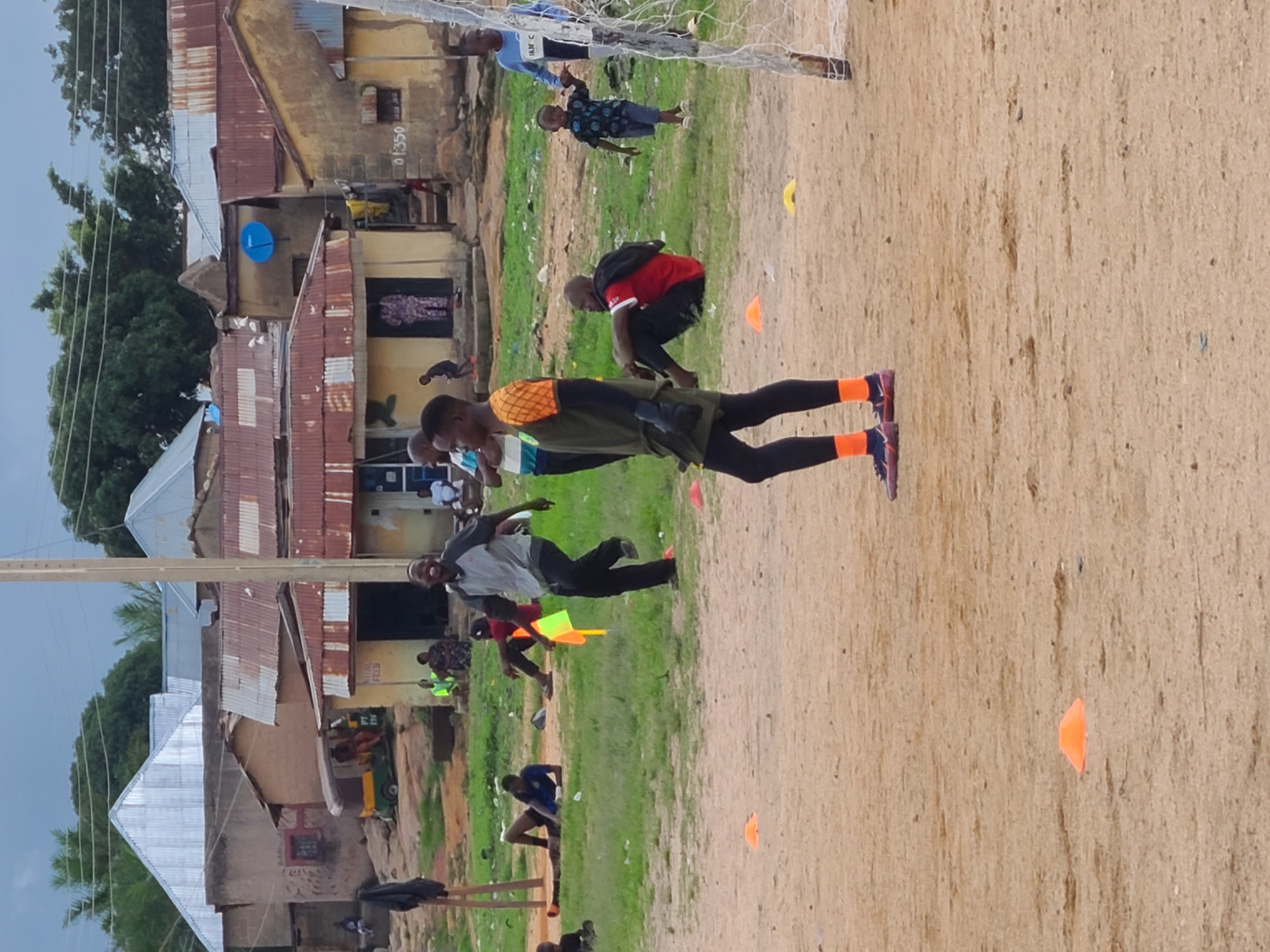 The image depicts an outdoor scene in a rural or semi-urban setting, likely in Jos, Nigeria. Several people are present, engaged in what appears to be a training or practice session, possibly for a sport like football, given the presence of a goalpost. The ground is a mixture of sandy soil and patchy green grass.
In the foreground, a young man in athletic wear, including orange socks and dark leggings, is the most prominent subject. He is captured in a dynamic pose, possibly mid-movement. Behind him, other individuals are scattered across the scene. Some are observing, while others seem to be participating in drills. One person in a red shirt and black shorts is crouching. Further back, a person in a blue shirt and shorts is near the goalpost, and another individual in a patterned shirt is also visible. Scattered on the ground are orange and yellow cones, commonly used for marking training areas.
The background features a cluster of buildings with corrugated iron roofs, suggesting a residential or community area. Some of these structures appear weathered. Trees are visible behind the buildings, and the sky is overcast, hinting at a cloudy or potentially humid day. There are no strong indicators of the exact time of day, but the lighting suggests it is daytime.
One building in the background has a visible number "01350" painted on its wall. The overall atmosphere seems active and communal, with a focus on athletic activity within a simple, everyday environment.