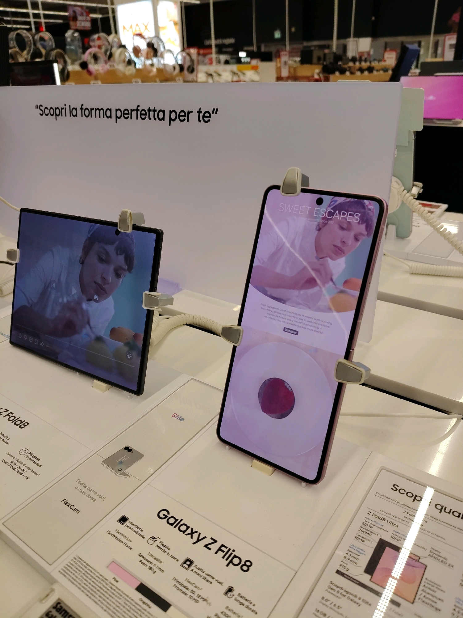 The image shows a retail display of Samsung Galaxy Z Fold 8 and Galaxy Z Flip 8 smartphones in a store. A white sign overhead reads "Scopri la forma perfetta per te" which translates to "Discover the perfect shape for you." The phones are displayed on white stands, with one phone open in a foldable position and another closed. There are price tags and product information labels on the display. The background shows shelves with various electronic devices, including headphones, and some lighting fixtures. The overall environment is a well-lit retail setting.