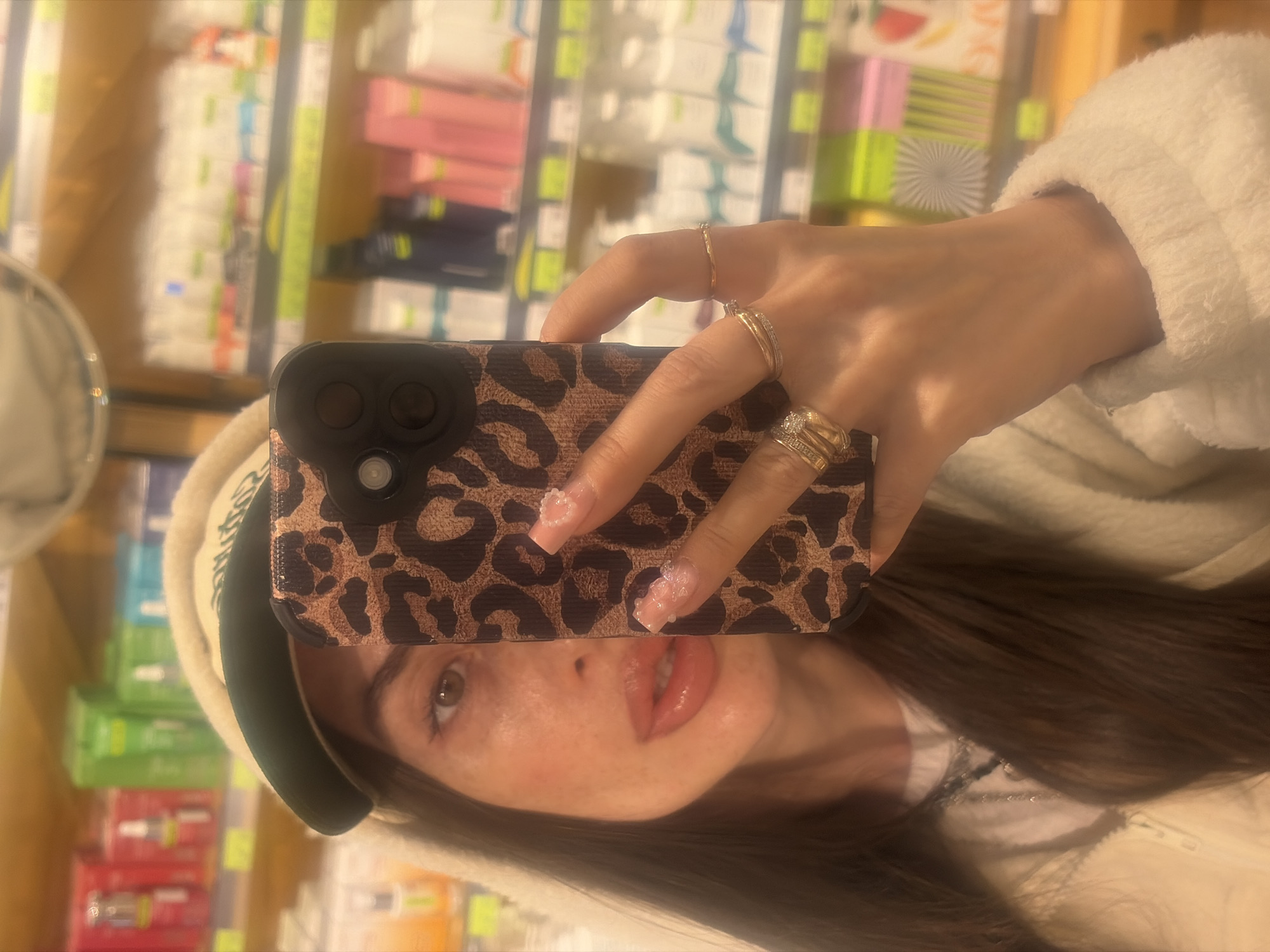 A close-up selfie is taken by a woman in what appears to be a retail store, possibly a pharmacy or beauty supply store, in Saint Petersburg, Russia. The woman is holding up her phone, which has a leopard print case, to capture the reflection in a mirror. Her right eye, nose, and lips are visible, along with her hand adorned with rings and detailed fingernails. The background is out of focus, showing shelves stocked with various products, suggesting a commercial environment. The lighting is warm, creating a soft, somewhat hazy effect.