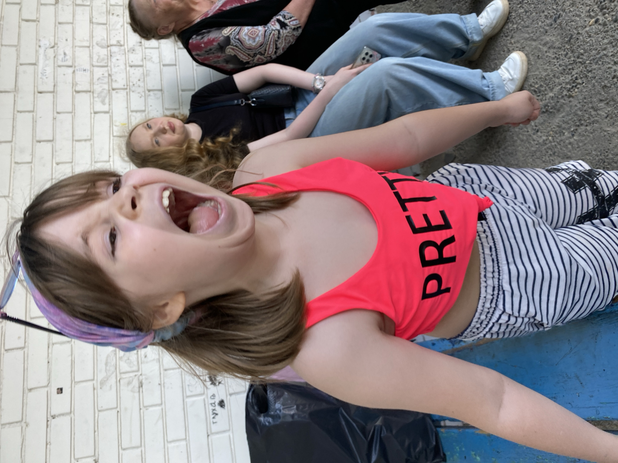 A young girl wearing a pink top with the word "PRETTY" printed vertically in black lettering, a purple headband, and striped pants is shouting. The background shows a light-colored brick wall. Behind the girl, two other women are sitting: one with blonde hair wearing a black top and jeans, and an older woman with gray hair wearing a patterned top and jeans. The setting appears to be an outdoor urban environment with a rough, grayish ground surface visible in the background. A black plastic bag is also present. Based on the information, it is believed to be in the city of Noviy, Russia.