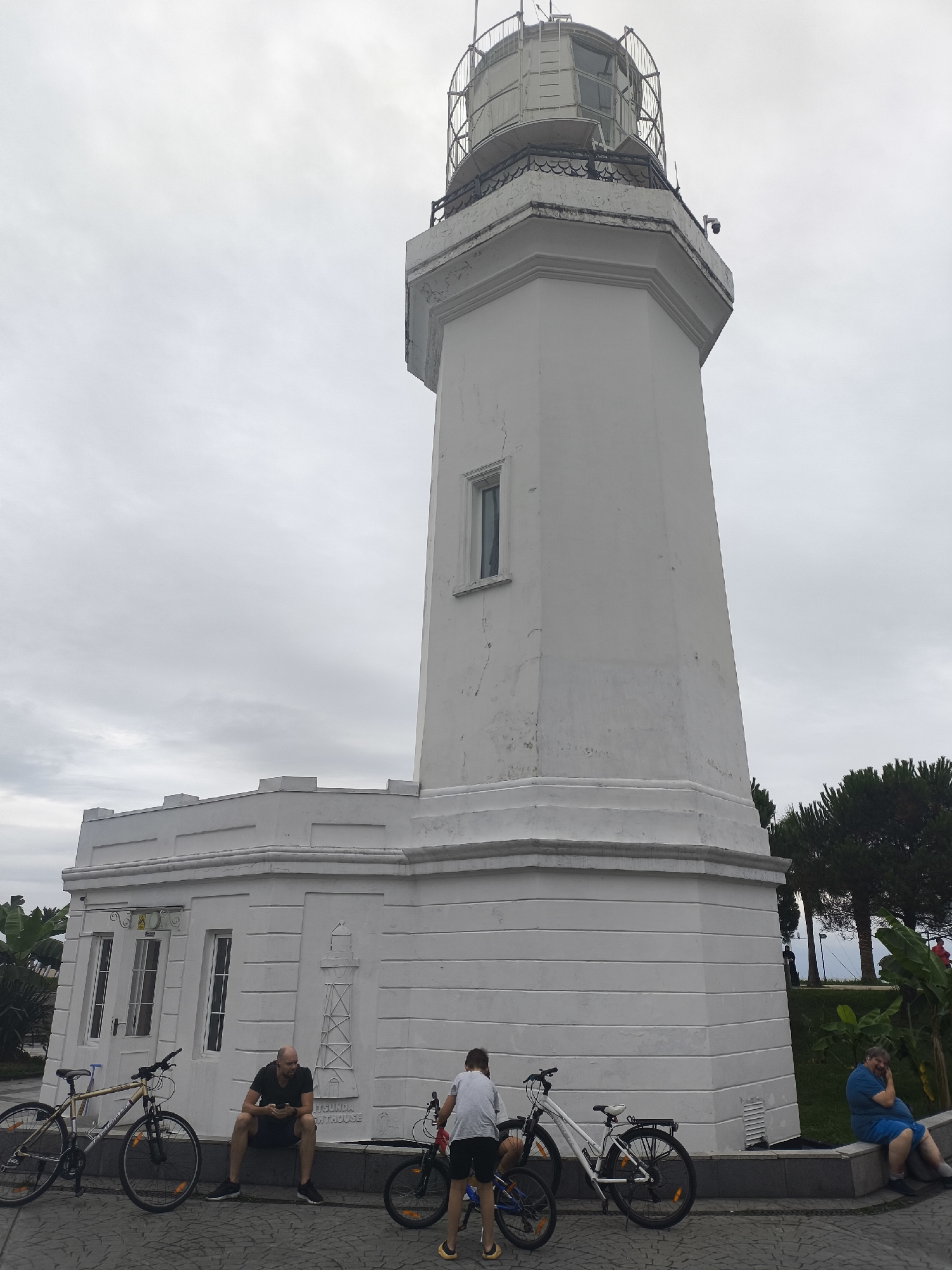 The image depicts the Batumi Lighthouse, a prominent landmark in Batumi, Georgia. The lighthouse is a tall, white, cylindrical structure with a lantern room at the top. In front of the lighthouse, there is a low-lying white building that appears to be part of the lighthouse complex. The ground is paved with cobblestones.
Several people are present in the scene. On the left side, a man is sitting on a ledge in front of the white building, looking at his phone. Two bicycles are parked nearby. In the center, a child is standing next to a white bicycle, and another man is visible in the background, also near a bicycle. On the right side, a person is sitting on a ledge, looking towards the lighthouse.
The sky is overcast, suggesting a cloudy day. Lush green trees and foliage are visible in the background, indicating a natural environment surrounding the lighthouse. The overall scene captures a moment of leisure and tourism at a historical site in Batumi.