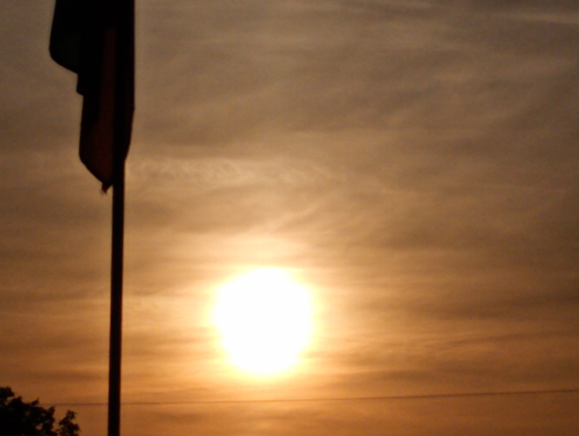 The image captures a sunset scene in Roza, Bulgaria. A flagpole, with a dark flag partially visible, stands in the left side of the frame. The sun appears as a bright, diffuse orb near the center of the image, creating an orange-yellow glow in the sky. The sky displays some cloud formations. The horizon is visible near the bottom of the frame with some trees on the left side.