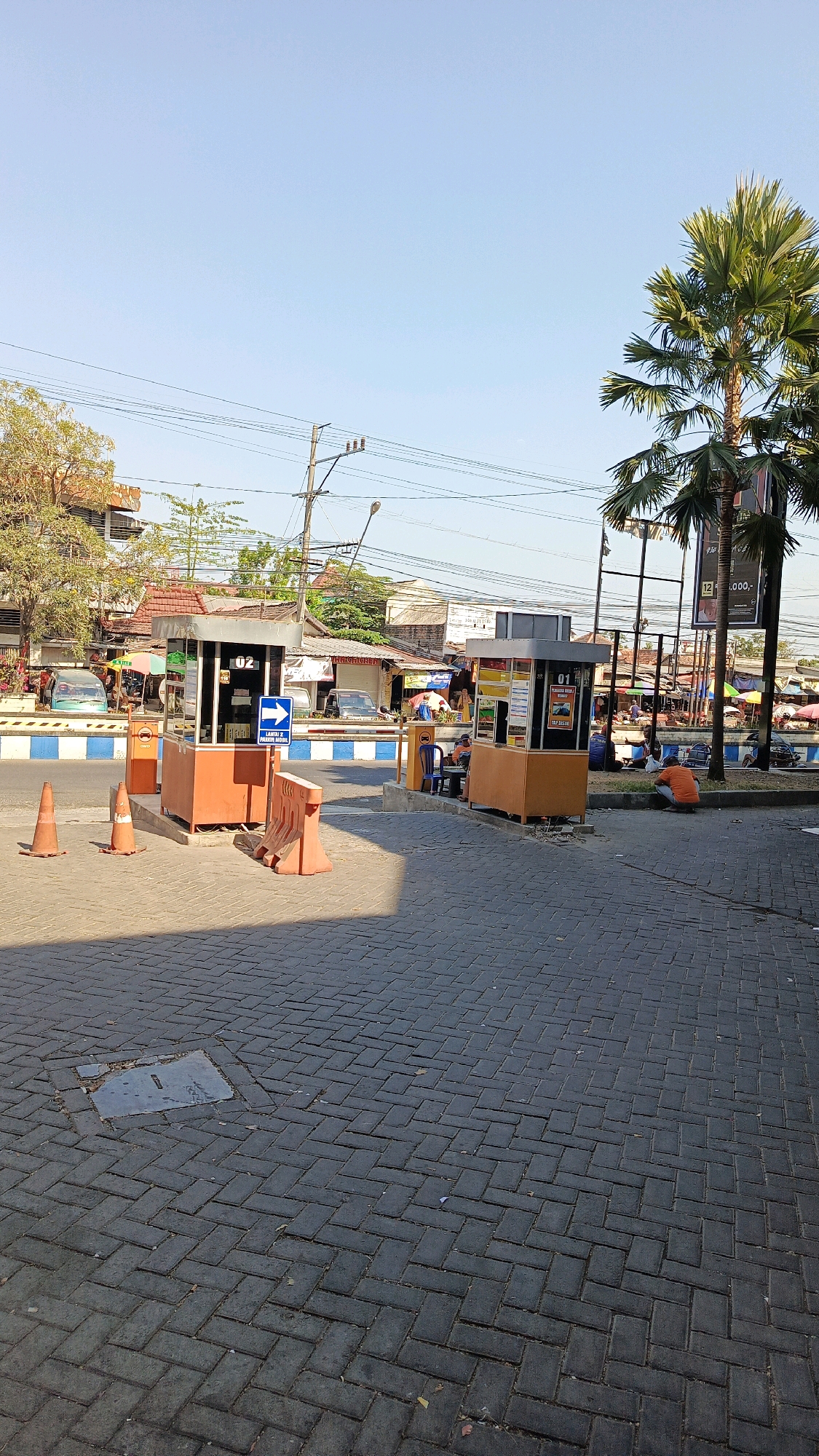 The image captures a street scene in Ponorogo, Indonesia, with a clear blue sky overhead. In the foreground, a paved area with interlocking gray bricks is visible, with a metal grate set into the pavement. Two orange security booths, likely for a toll or parking area, are positioned on the right side of the street. The booth on the left has a blue arrow sign pointing to the right and the text "MASUK 2 MOBIL." The booth on the right has a sign with the number "01." A blue barrier is present in front of the booths.
Several orange traffic cones are placed on the left side of the paved area. In the background, palm trees and other vegetation are visible, along with buildings and a street lined with stalls or shops. There are a few cars parked or driving in the background. A person in an orange shirt is seen crouching on the right side of the image, near the booths. Another person is seated in a blue chair in front of the booth on the right. The overall impression is a public space during the daytime.