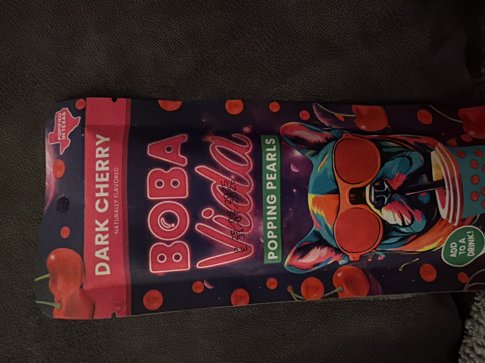 The image shows a product package for "Boba Vida Popping Pearls" in Dark Cherry flavor. The package is dark blue with a pink border and features stylized illustrations of cherries and a colorful French bulldog wearing sunglasses. The text "DARK CHERRY NATURALLY FLAVORED" is prominently displayed. In the top right corner, there is a silhouette of Texas with the text "POPFIED IN TEXAS". Below the brand name, there is a batch and expiry date, which appears to be P 25/08/2025 and E 31/01/2026. Towards the bottom, there is text that reads "ADD TO A DRINK!". The package is lying on a dark, textured surface. There are no people or identifiable city locations present in the image.