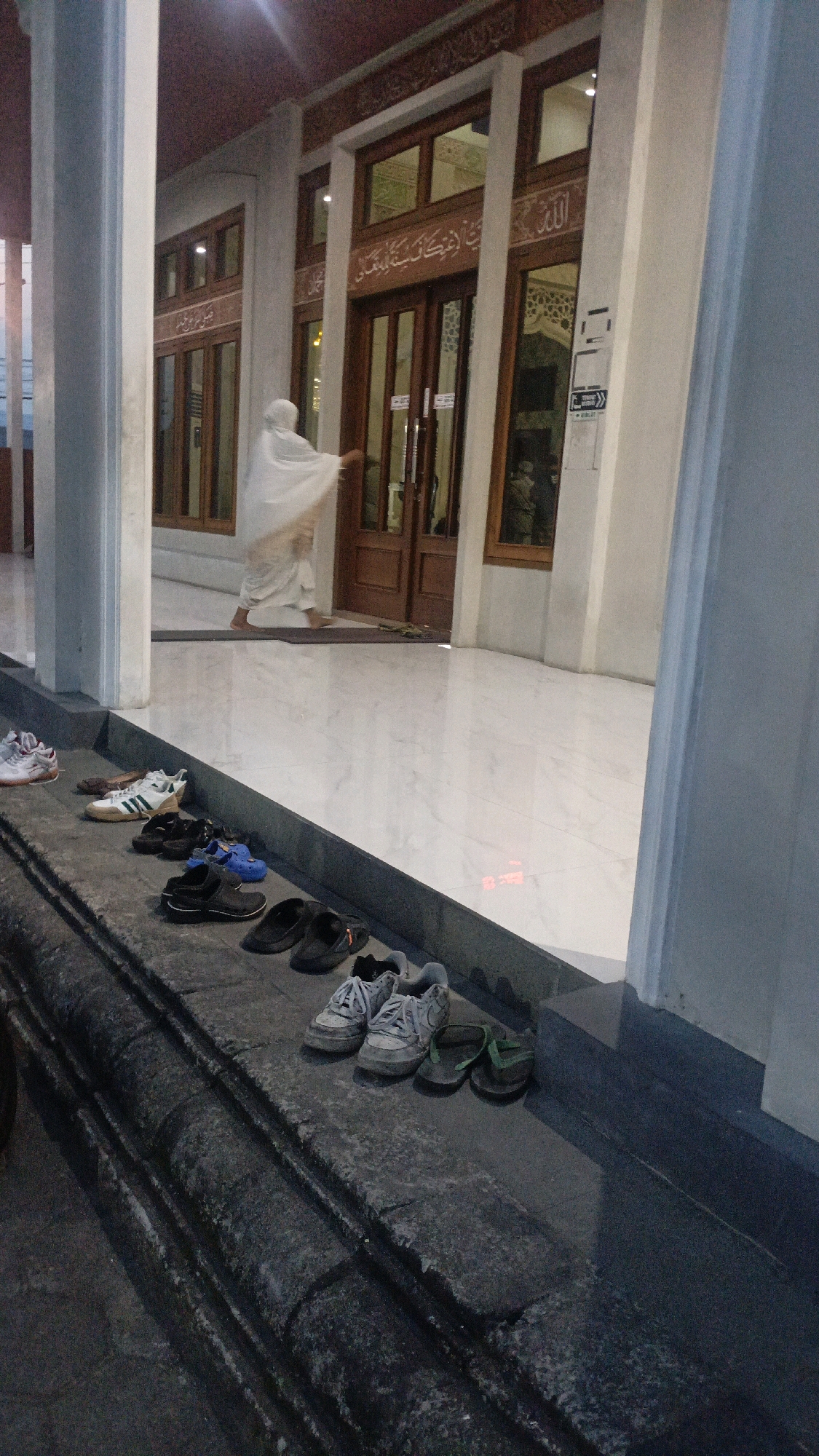 The image depicts the entrance of a building, possibly a mosque, with a person in a white head covering and long robe walking towards the doors. In the foreground, on a stone ledge, are several pairs of shoes, including sneakers, sandals, and flip-flops. The floor inside the building is made of light-colored marble. The architecture suggests it is a public building, likely in a place of worship. The lighting indicates it is either early morning or late evening. The location is specified as Ngaru Aru, Indonesia.