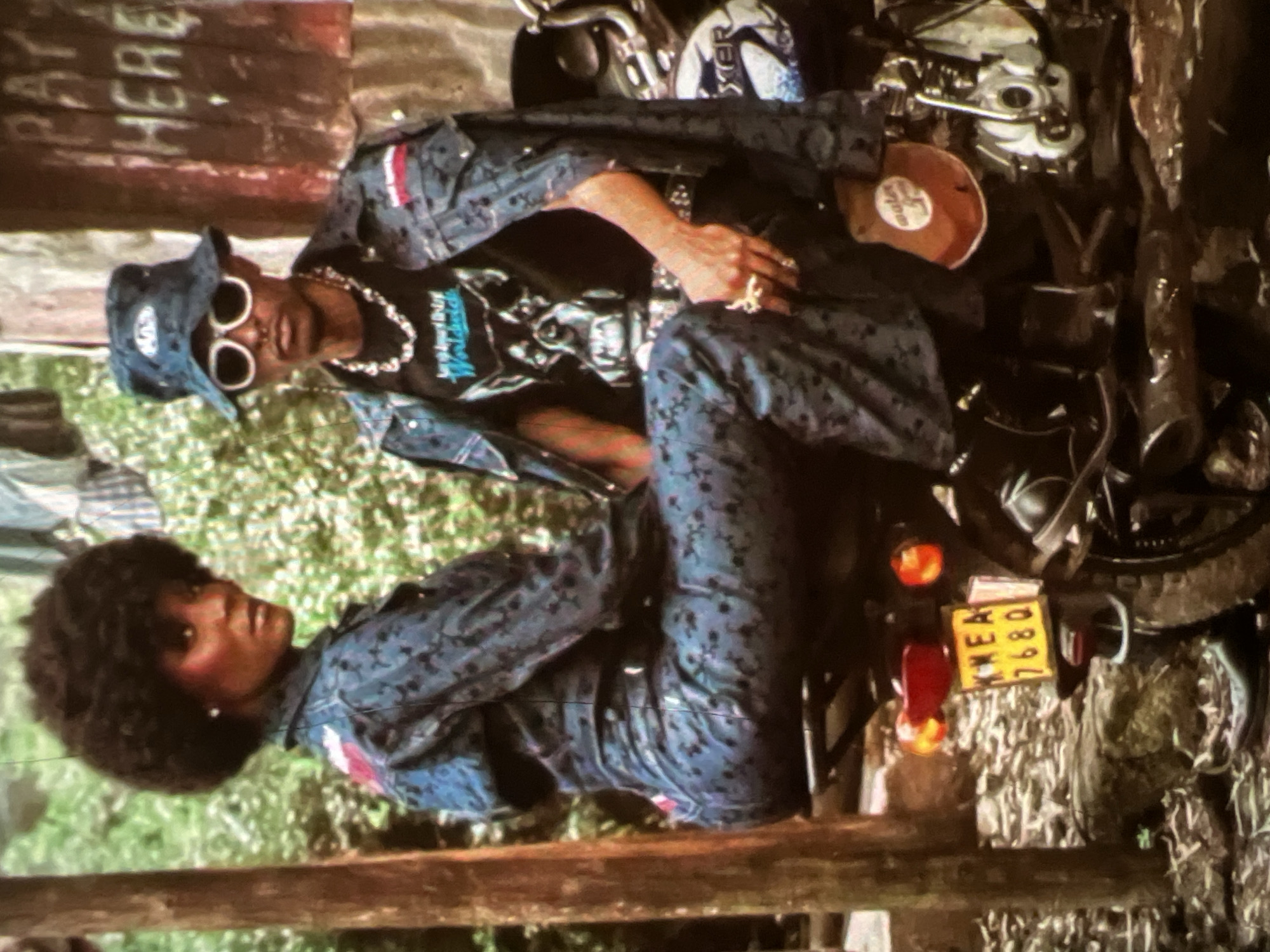 The image depicts a man and a woman posing on a motorcycle, outdoors in a possibly informal setting within Lagos, Nigeria. The woman, with an afro hairstyle and dark skin, is seated on the back of the motorcycle, looking towards the viewer. She is wearing a blue patterned jumpsuit and a red patch on her left shoulder. The man is seated on the front of the motorcycle, facing forward with his head turned slightly towards the viewer. He has dark skin, is wearing a blue patterned jacket over a black t-shirt with a graphic print, a bucket hat, and distinctive white-rimmed sunglasses. He is also adorned with several necklaces and rings. The motorcycle's license plate reads "KNEA 7680". In the background, there is a wooden post on the left, a wall with writing that includes "PAY HERE" on the right, and foliage suggesting an outdoor environment. The overall impression is a fashion-oriented or artistic portrayal.