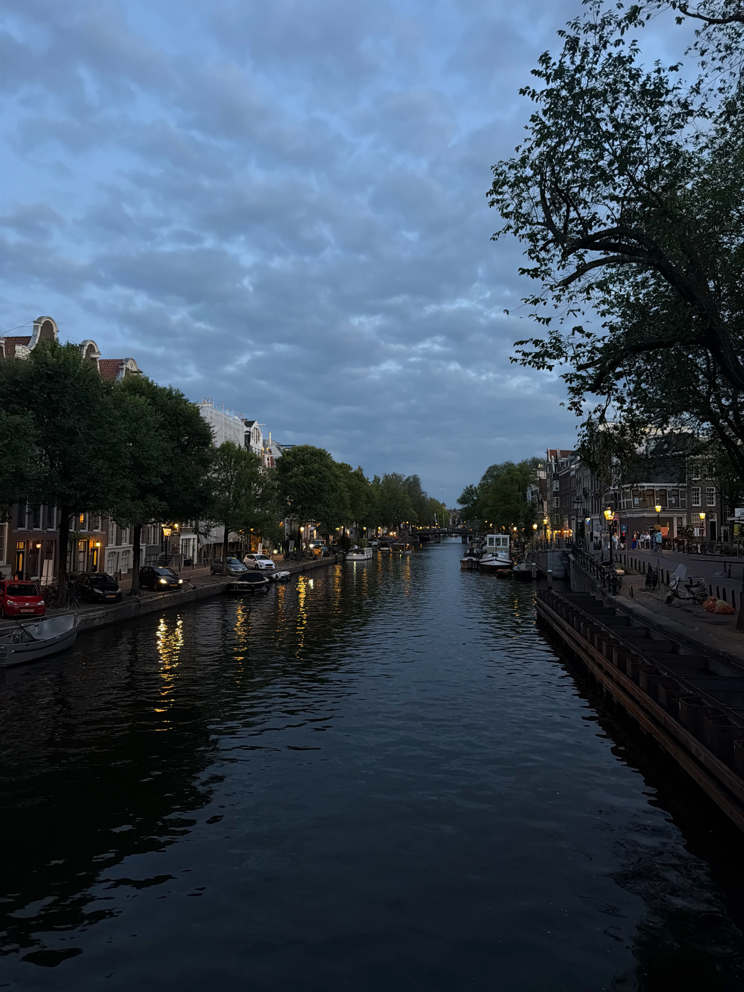 A canal runs through Amsterdam, Netherlands, lined with historic buildings and trees. The sky is overcast with dramatic clouds, and the water reflects the lights from the buildings and streetlamps. Several boats are moored along the canal, and a few cars are parked on the street. A small group of people can be seen walking on the sidewalk. The scene appears to be captured at dusk, creating a moody and atmospheric ambiance.