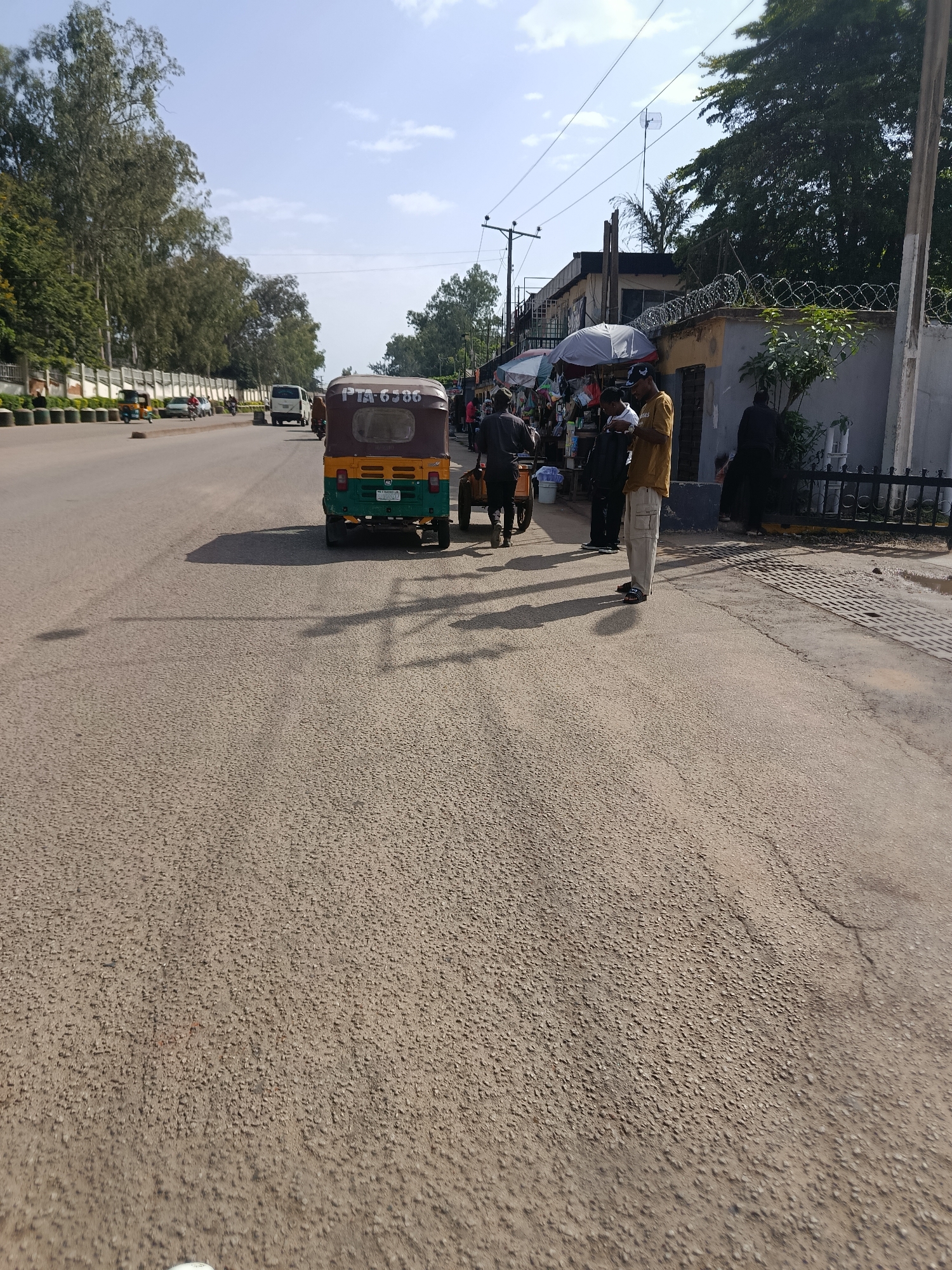 The image depicts a street scene in Jos, Nigeria, on a clear day. A three-wheeled auto-rickshaw, or "Keke Napep," with license plate "PTA 6586" is parked on the right side of the road. In front of a building, several individuals are gathered near market stalls displaying various goods under umbrellas. Some people are standing, one person is pushing a cart, and another is looking at a mobile phone. The building behind the market appears to be a commercial establishment with a fence topped with barbed wire. The road surface is asphalt, and trees line the left side of the street, with a wall and streetlights visible. The sky is blue with scattered clouds.