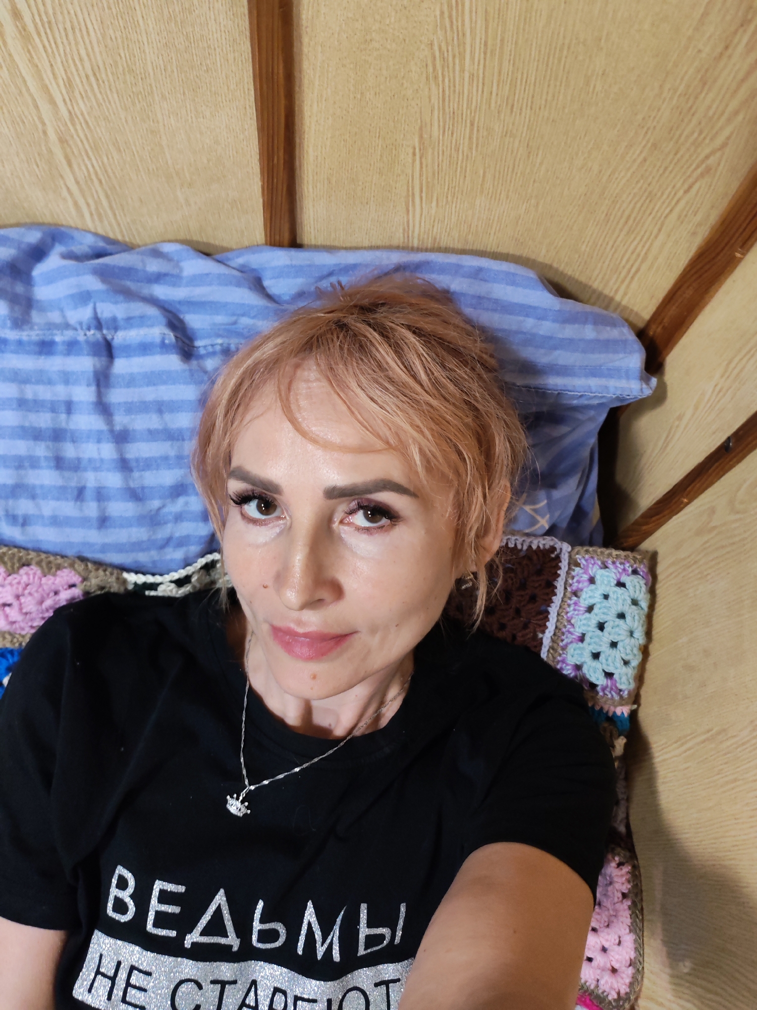 In a dimly lit room, a woman with peach-colored hair and makeup is lying on a bed. She is wearing a black t-shirt with silver Cyrillic lettering and a delicate silver necklace. The bed is covered with a blue striped sheet and a crocheted blanket featuring squares of pink, brown, and light blue. The wall behind her is made of light-colored wooden planks. The overall impression is one of a domestic setting, possibly a bedroom. The specific location in Bol'shie Mkhi, Russia, is not discernible from the image itself.