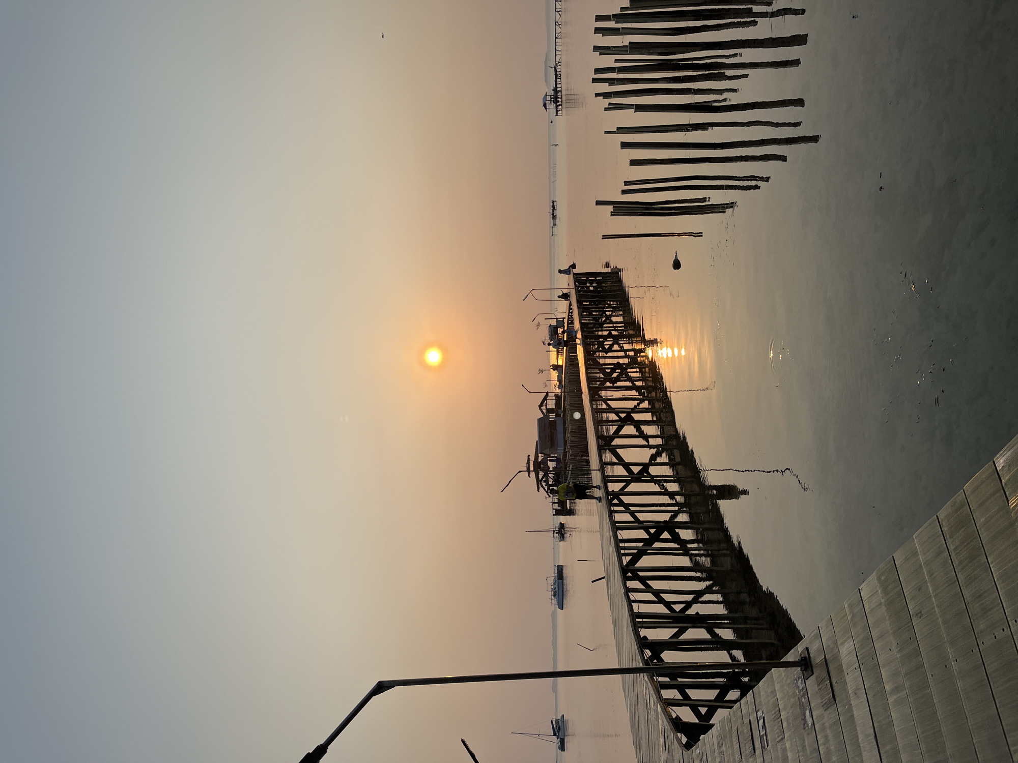 An outdoor, eye-level, vertical shot captures a serene sunset over a calm body of water. The foreground features a wooden pier extending into the water, its planks weathered and reflecting the warm hues of the sky. Several wooden posts stand vertically in the water to the right of the pier, creating a rhythmic pattern. In the mid-ground, the pier continues, leading to a small structure with a roof and several boats docked nearby. The sun, a bright orb, is visible in the upper center of the frame, casting a warm glow and a shimmering reflection on the water's surface. The sky transitions from a pale orange near the horizon to a lighter blue towards the top. A few distant boats and structures are discernible on the horizon. The overall atmosphere is peaceful and tranquil, indicative of a coastal location at dusk. This scene is located in Kemloko, Indonesia.